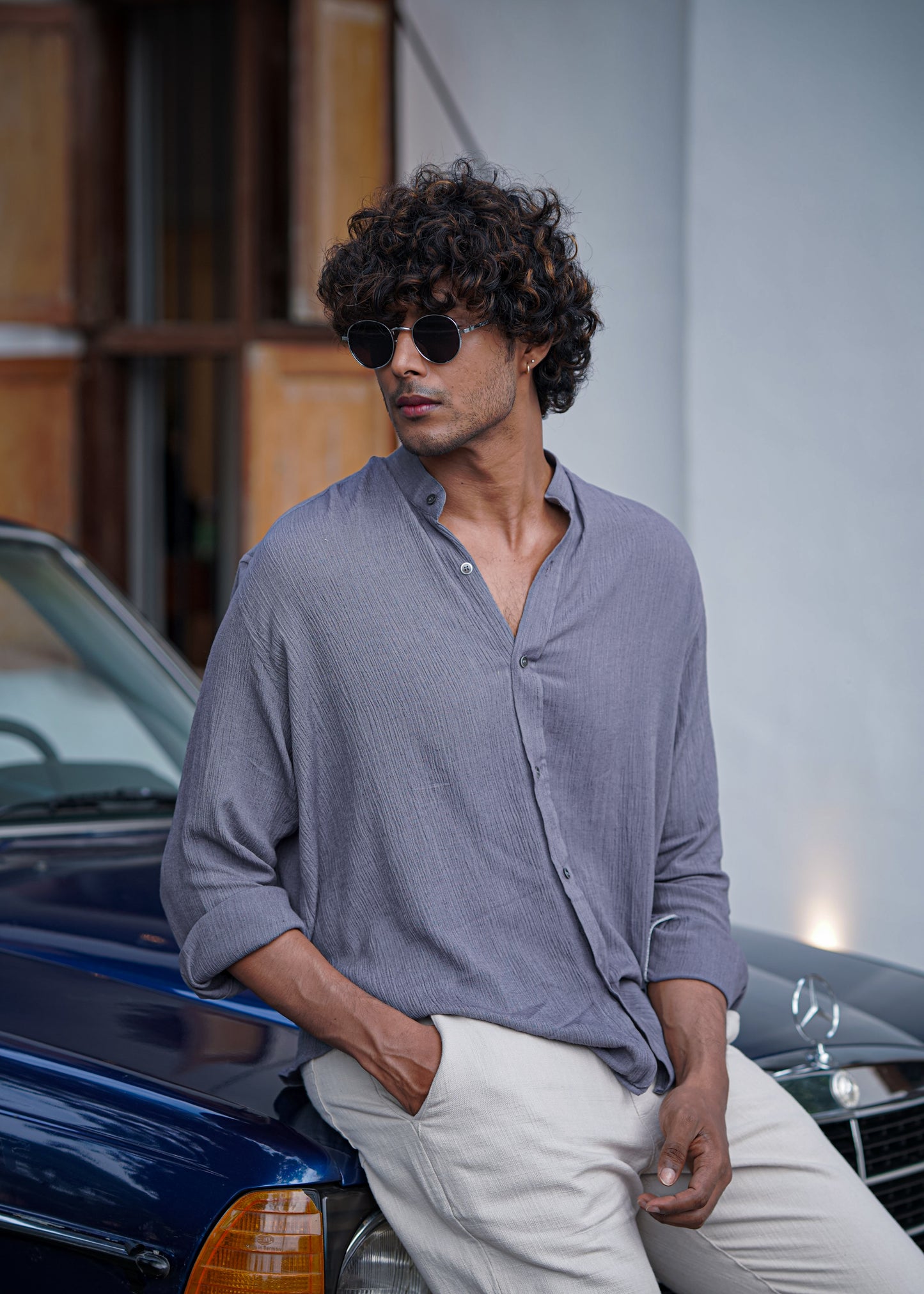 Mod Maven Lilac Grey Crushed Cotton Full Sleeve Shirt paired with Off-White Comfortable Fit Pants. Model sitting on a vintage dark blue car, wearing shades, showcasing a chic street style look.