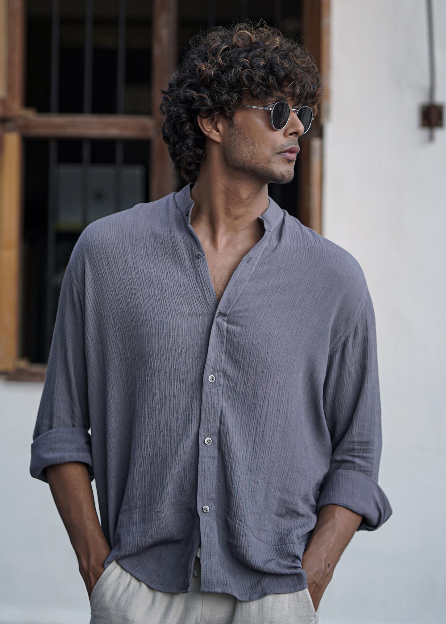 Mod Maven Lilac Grey Crushed Cotton Full Sleeve Shirt paired with Off-White Comfortable Fit Pants. Zoomed in image of Model standing in front of a wooden window wearing shades, showcasing a chic street style look.