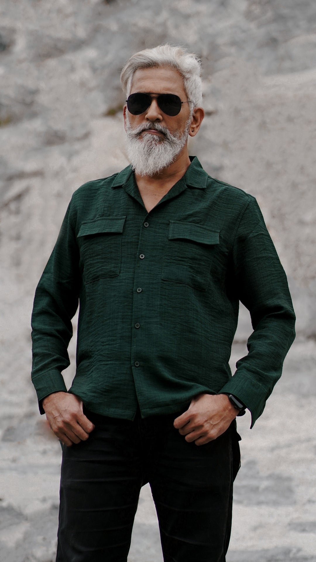 A distinguished older man with a gray beard and stylishly combed gray hair is wearing an army green, double-pocket, full-sleeve shirt from Mod Maven. He pairs the shirt with dark trousers and accessorizes with black aviator sunglasses and a sleek black wristwatch, standing confidently against a textured, natural background. The overall look exudes modern sophistication and timeless style.