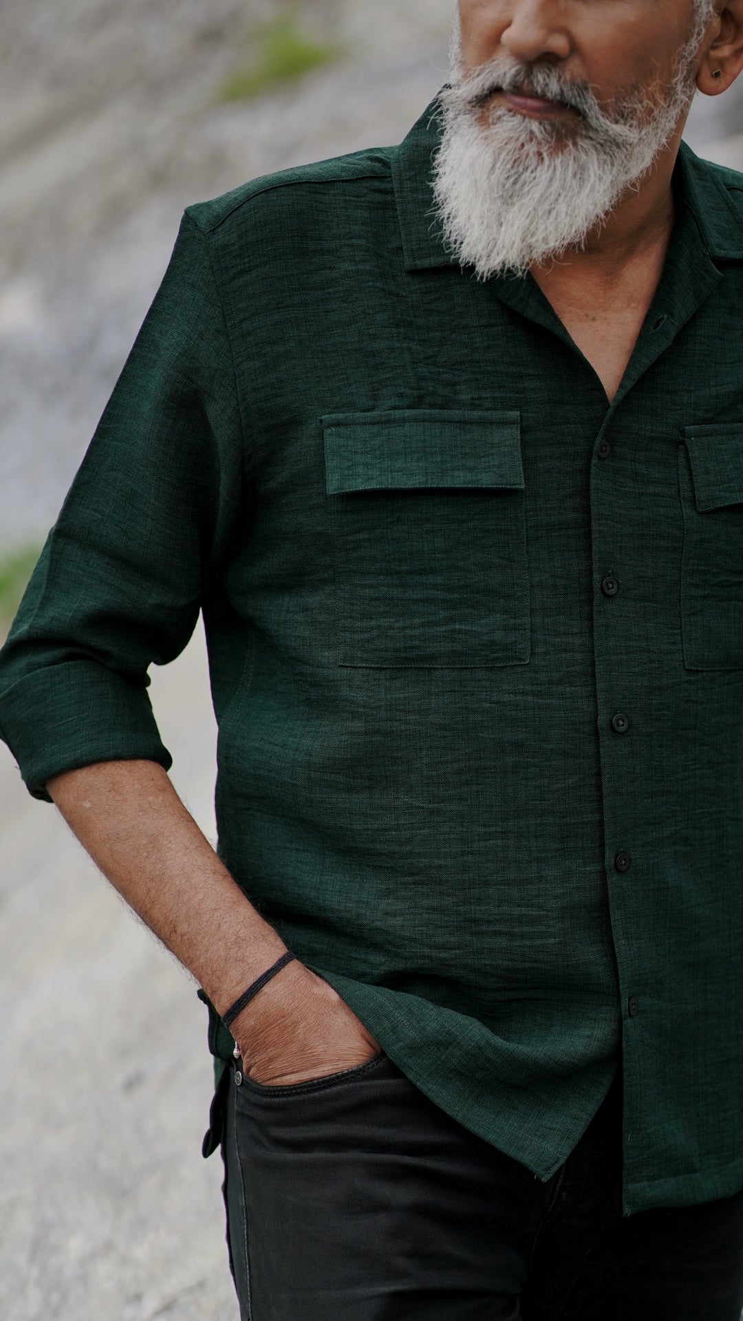 A distinguished older man with a gray beard and stylishly combed gray hair is wearing an army green, double-pocket, full-sleeve shirt from Mod Maven. He pairs the shirt with dark trousers and accessorizes with a sleek black wristwatch, standing confidently against a textured, natural background. The picture focuses on the collar and pocket detail