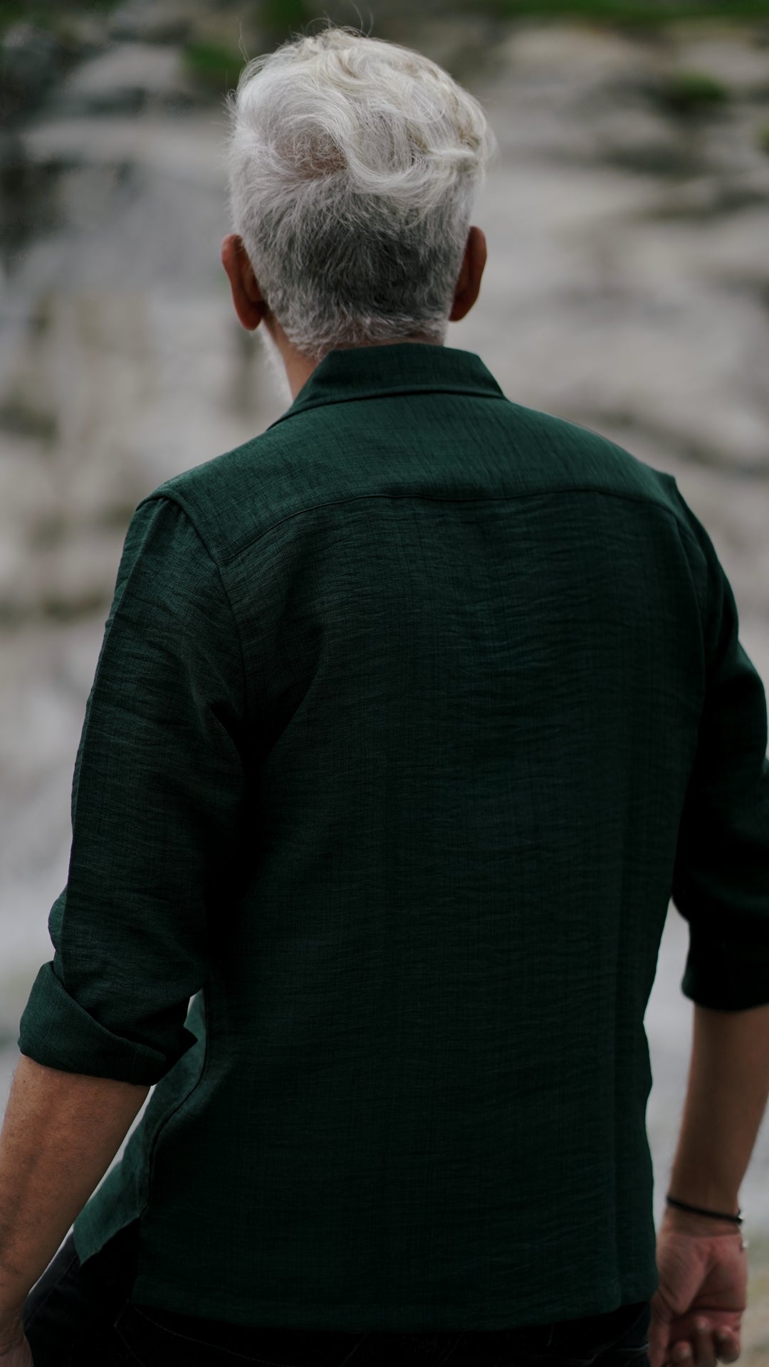 Back view of a stylish older man with a neatly combed gray hair is showcasing an army green, double-pocket, full-sleeve shirt from Mod Maven. Set against a rocky backdrop conveying a sense of adventure and elegance. His outfit is completed with black trousers, black shoes, emphasizing his effortlessly cool and sophisticated style.