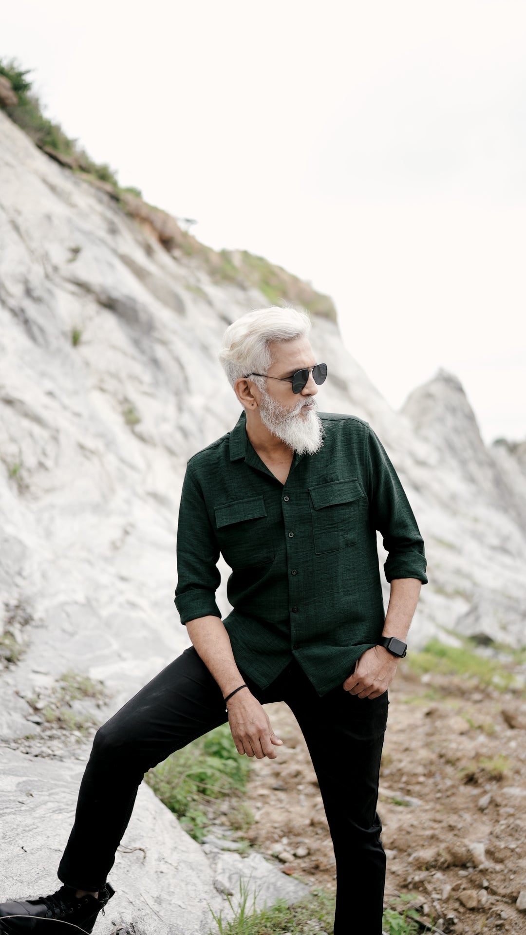 A stylish older man with a gray beard and neatly combed gray hair is showcasing an army green, double-pocket, full-sleeve shirt from Mod Maven. Set against a rocky backdrop, he poses with one leg resting on a rock, conveying a sense of adventure and elegance. His outfit is completed with black trousers, black shoes, and aviator sunglasses, emphasizing his effortlessly cool and sophisticated style.