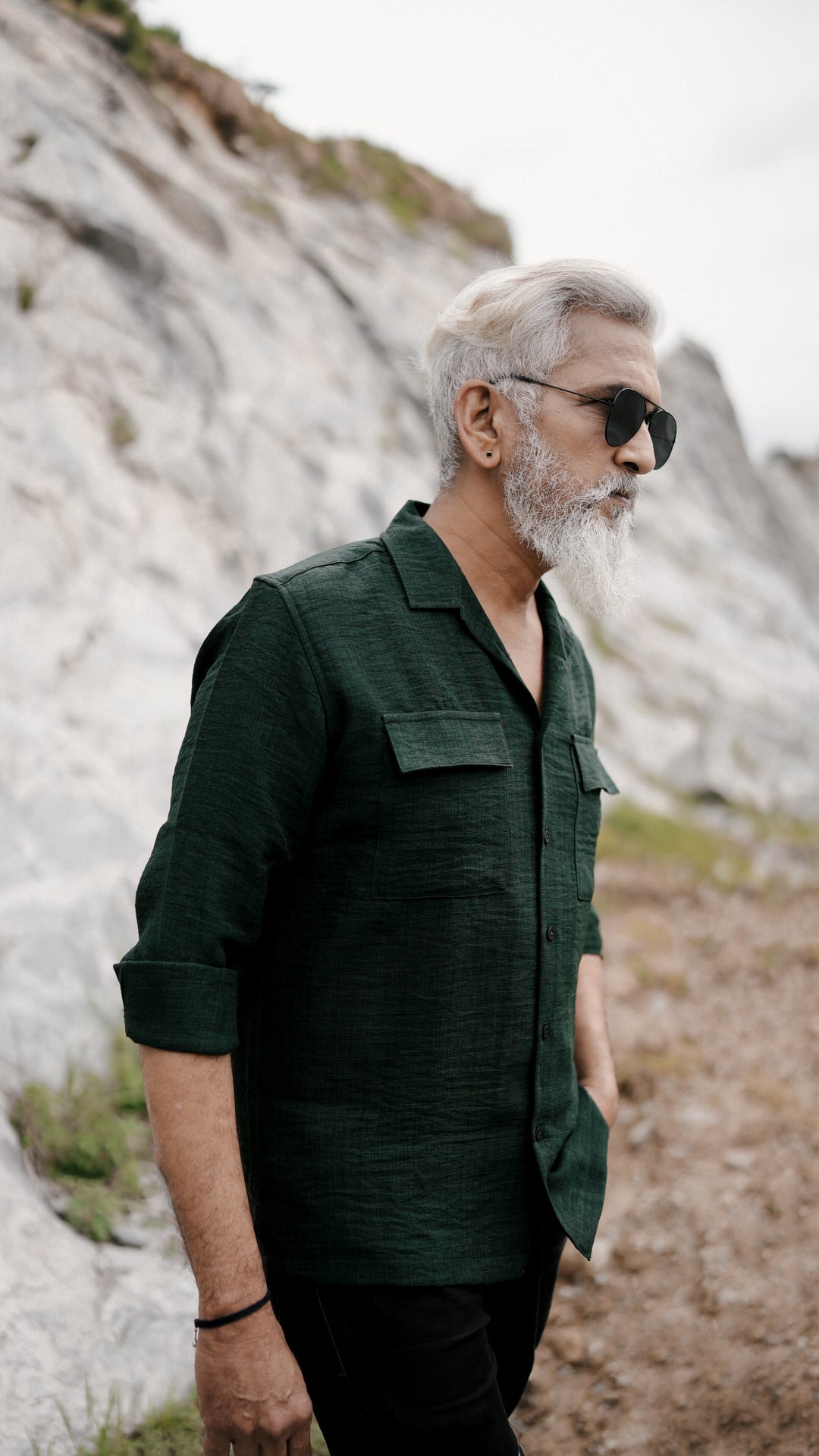 A stylish older male model with a gray beard and neatly combed gray hair is showcasing an army green, double-pocket, full-sleeve shirt from Mod Maven. Set against a rocky backdrop, he poses conveying a sense of adventure and elegance. His outfit is completed with black trousers, black shoes, and aviator sunglasses, emphasizing his effortlessly cool and sophisticated style.