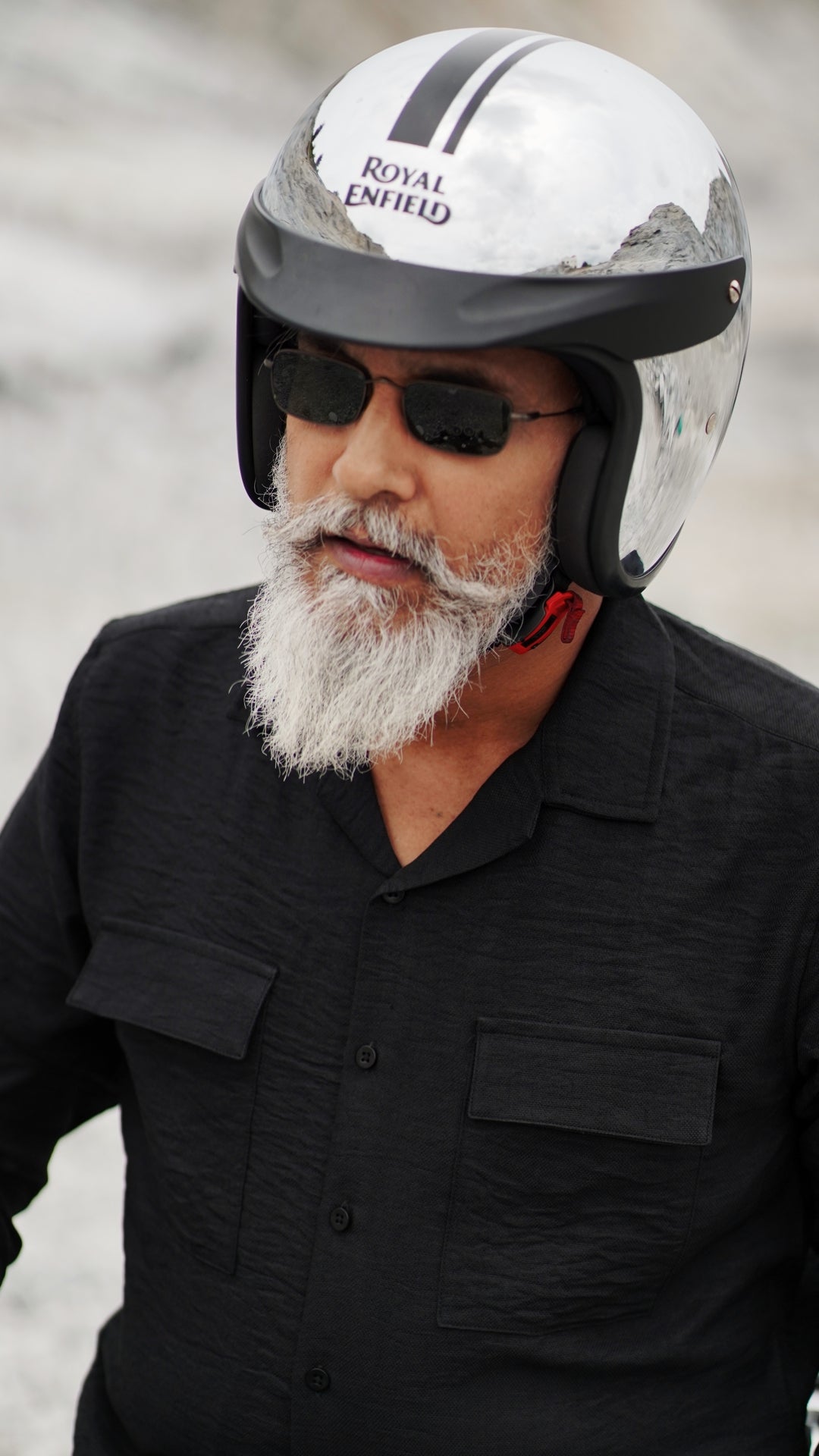 An older male model with a gray beard is wearing a black, double-pocket, full-sleeve shirt from Mod Maven. He pairs the shirt with dark trousers and accessorizes with black sunglasses, sitting on a Royal Enfield Continental Gt wearing a metallic finish helmet, confidently against a textured, natural background. The overall look exudes modern sophistication and timeless style.