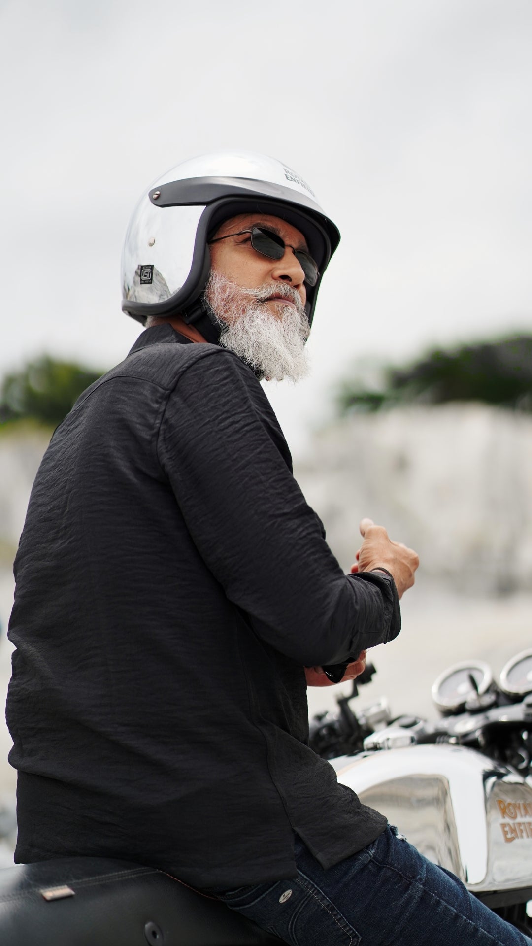 An older male model with a gray beard is wearing a black, double-pocket, full-sleeve shirt from Mod Maven. He pairs the shirt with dark trousers and accessorizes with black sunglasses, sitting on a Royal Enfield Continental Gt wearing a metallic finish helmet, confidently against a textured, natural background. The overall look exudes modern sophistication and timeless style.