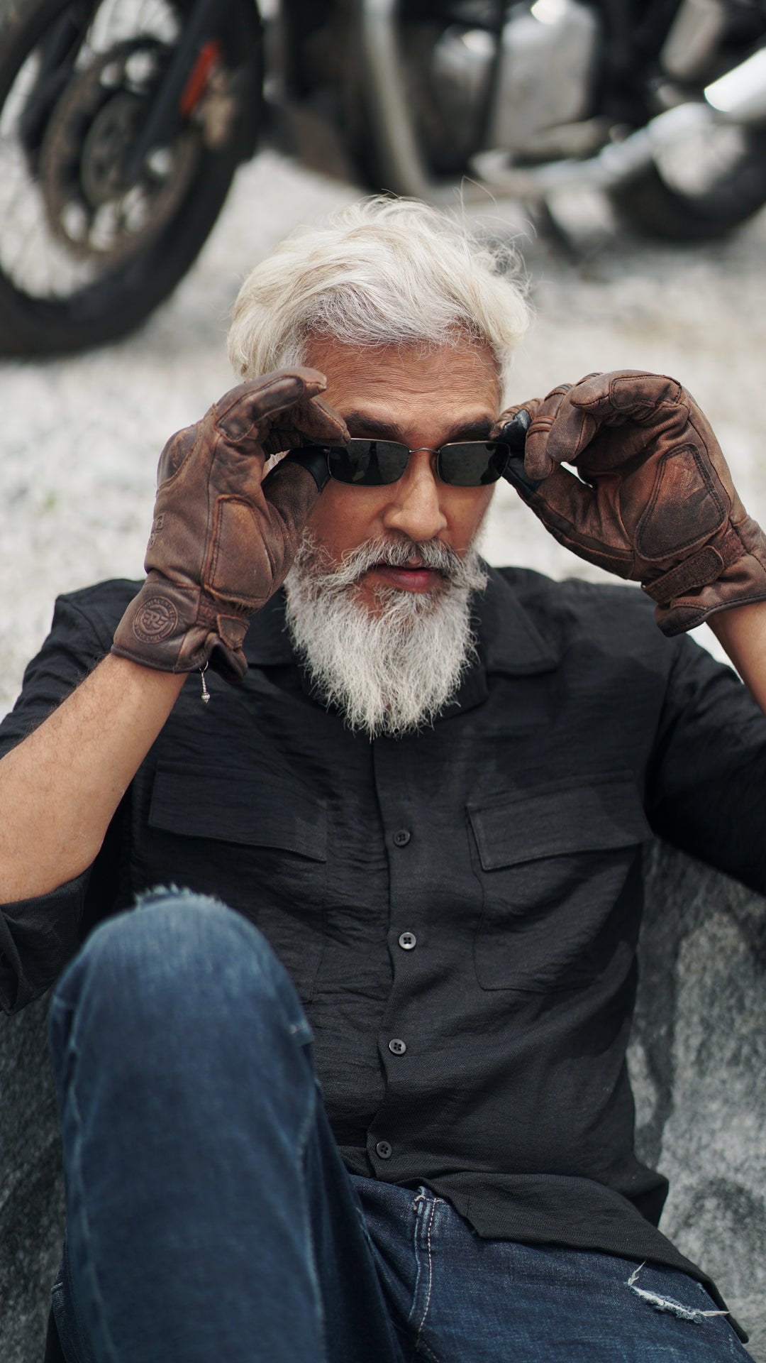 An older male model with a gray beard is wearing a black, double-pocket, full-sleeve shirt from Mod Maven. He pairs the shirt with dark trousers and accessorizes with black vintage sunglasses and gloves, sitting on a rock confidently against a textured, natural background. The overall look exudes modern sophistication and timeless style.