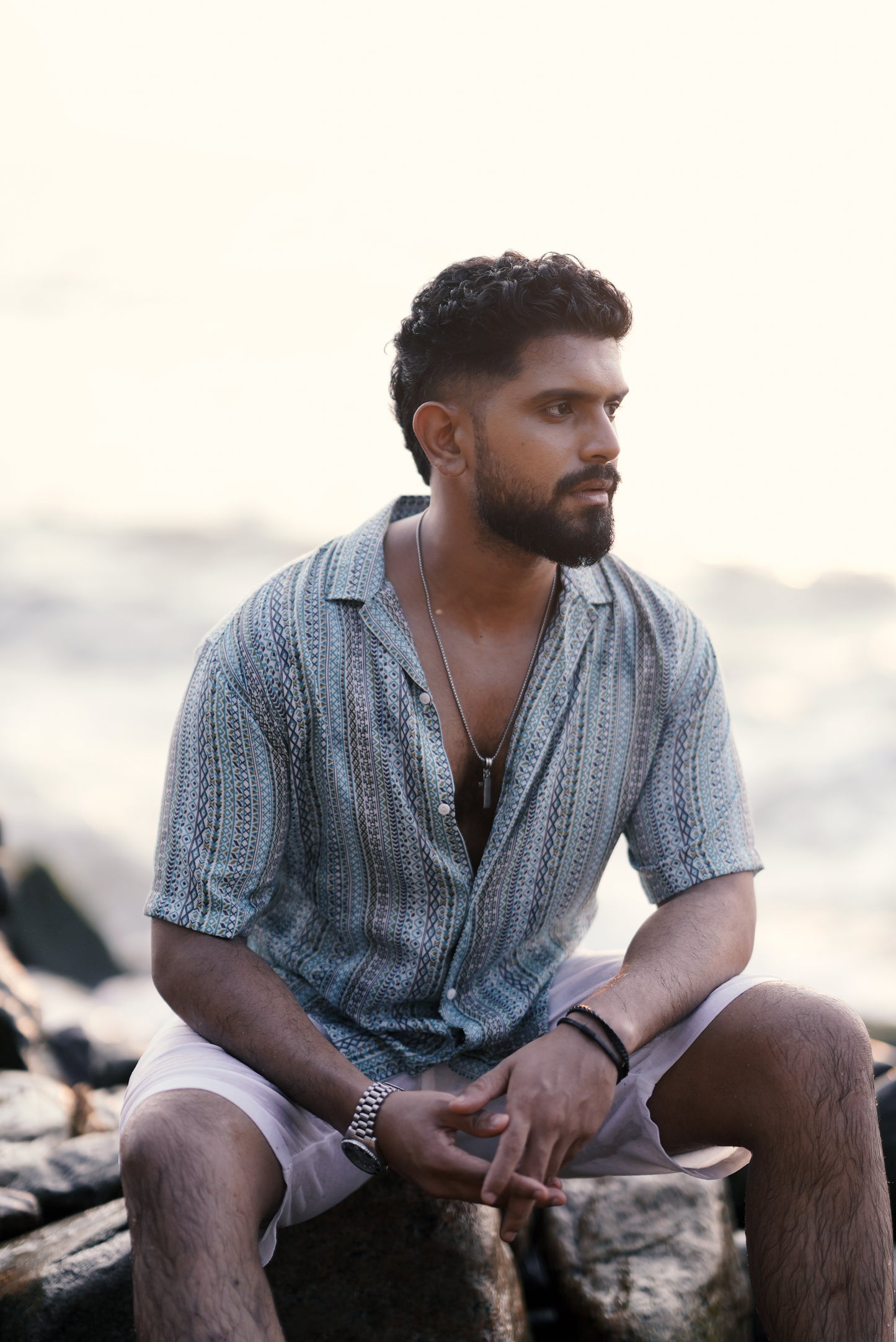 A model wearing a light green and aqua blue boho-inspired half-sleeve shirt with intricate patterns from Mod Mavens' menswear collection, captured sitting. He pairs the shirt with white linen shorts and stands on a rocky beach, turning slightly with a confident look.