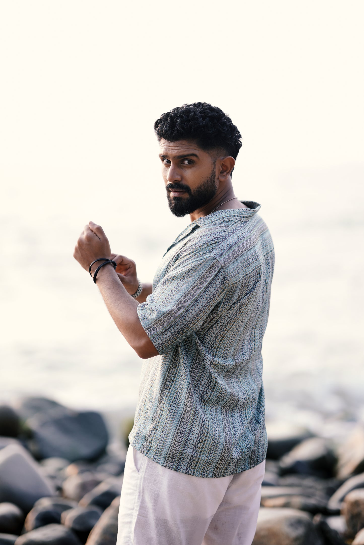 A modal wearing a light green and aqua blue boho-inspired half-sleeve shirt with intricate patterns from Mod Mavens' menswear collection, captured from the side. He pairs the shirt with white linen pants and stands on a rocky beach, turning slightly with a confident look.