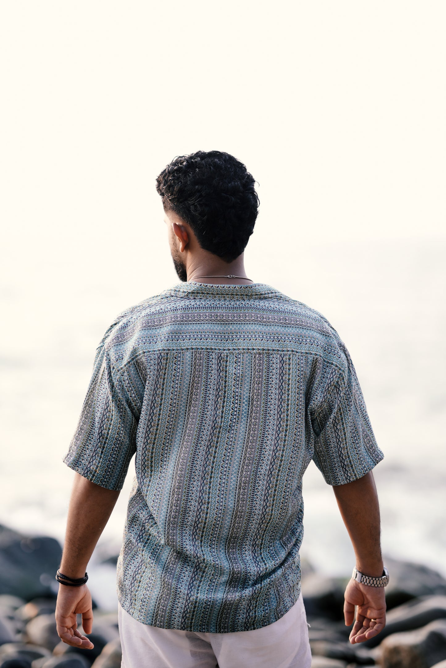 A back view of a modal wearing a light green and aqua blue boho-patterned half-sleeve shirt from Mod Mavens' menswear collection. The detailed pattern and relaxed fit are highlighted as he stands on a rocky shoreline, reflecting effortless coastal style.