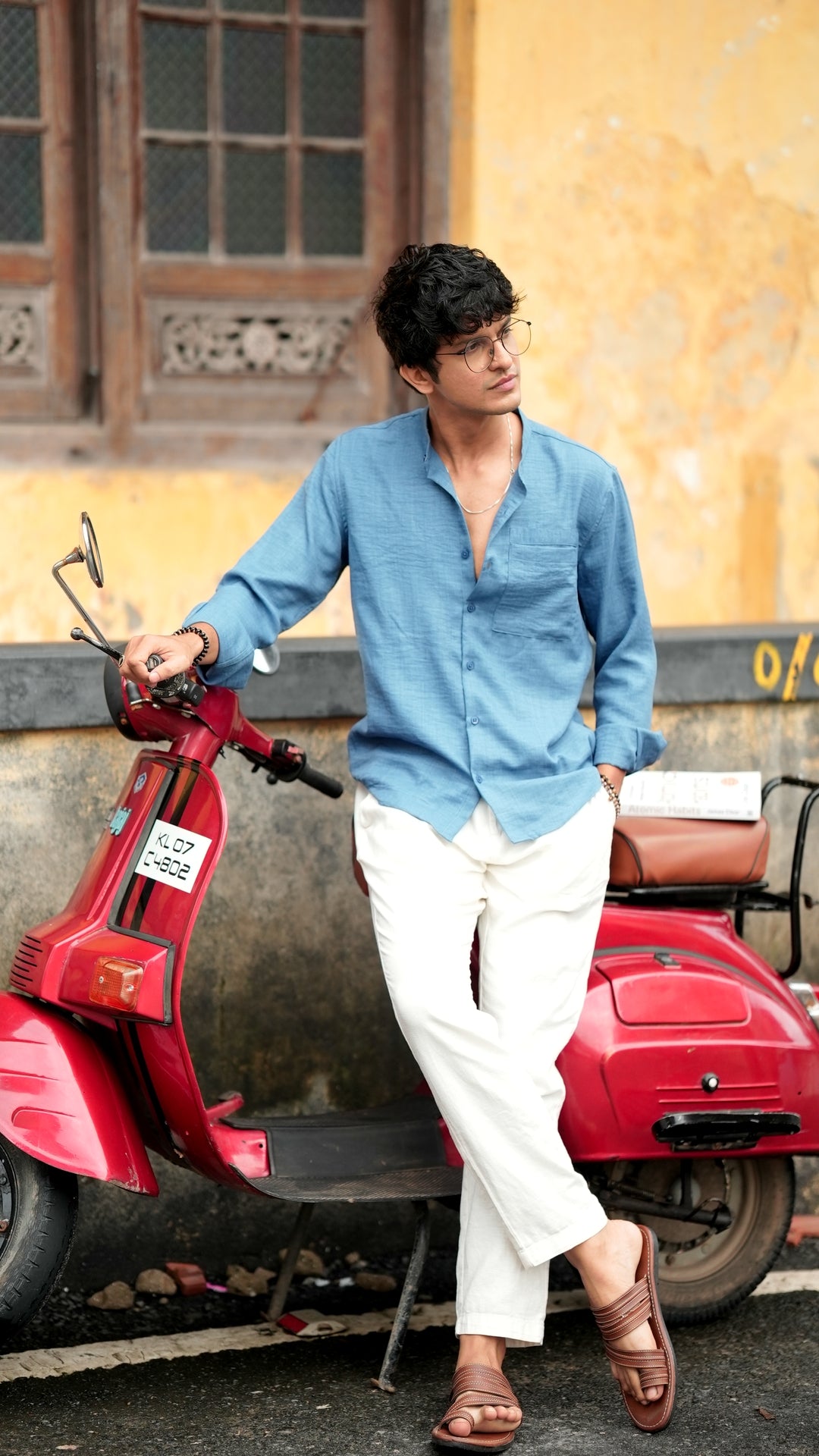 A male model sitting on a Chetak, wearing a denim blue, slim-fit, full-sleeve shirt from Mod Maven. He has short, tousled black hair, and wears round, reading glasses. The shirt has a soft texture with matching blue buttons. He is standing in front of a neutral-toned wall, exuding a casual yet refined look.