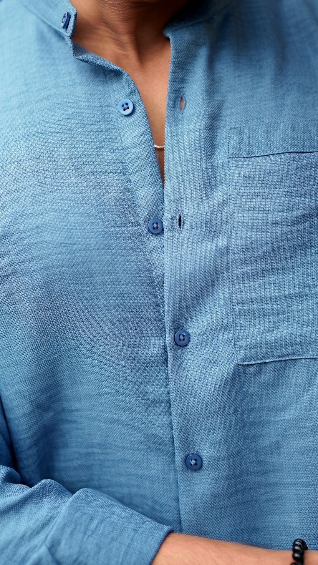 Close-up view of a male model wearing a denim blue, slim-fit, full-sleeve shirt from Mod Maven. The shirt is buttoned down with matching blue buttons, showing a sleek, minimalistic design. He is accessorized with a thin silver chain, adding a touch of elegance to the outfit. The background features a subtle, neutral wall that complements the soft tones of the shirt.