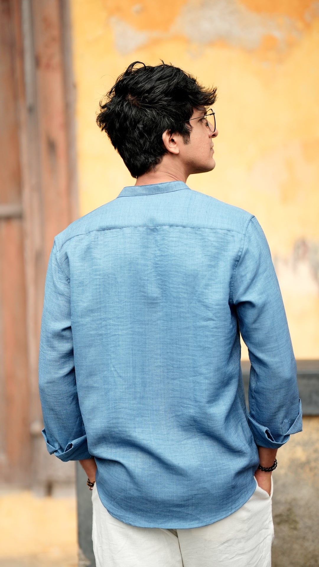 Back view of a male model wearing a denim blue, slim-fit, full-sleeve shirt from Mod Maven. He has short, tousled black hair, and wears round, reading glasses. The shirt has a soft texture with matching blue buttons. He is standing in front of a neutral-toned wall, exuding a casual yet refined look.