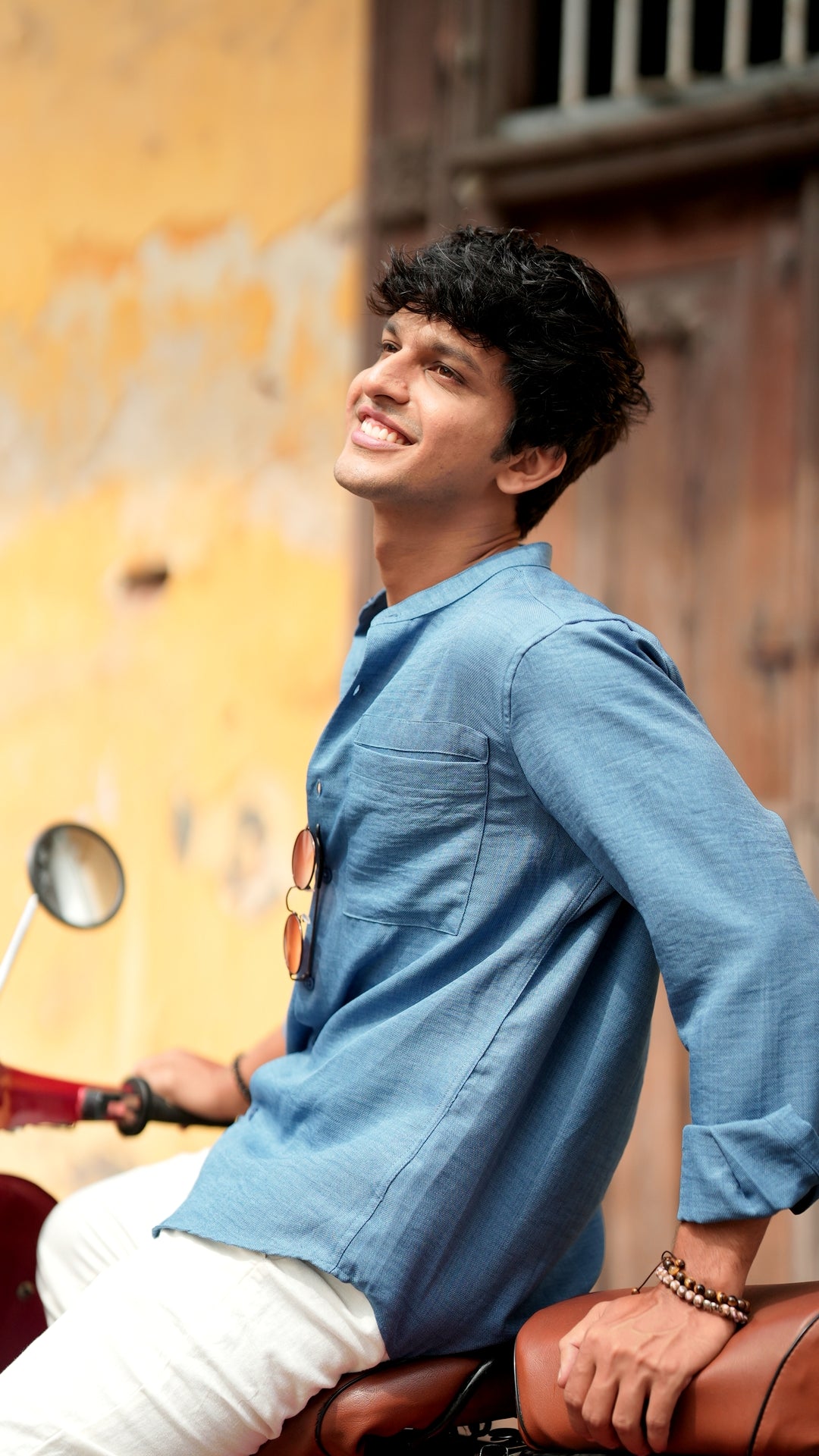 Side view of a male model sitting on a Chetak, wearing a denim blue, slim-fit, full-sleeve shirt from Mod Maven. He has short, tousled black hair, and wears round, reading glasses. The shirt has a soft texture with matching blue buttons. He is standing in front of a neutral-toned wall, exuding a casual yet refined look.