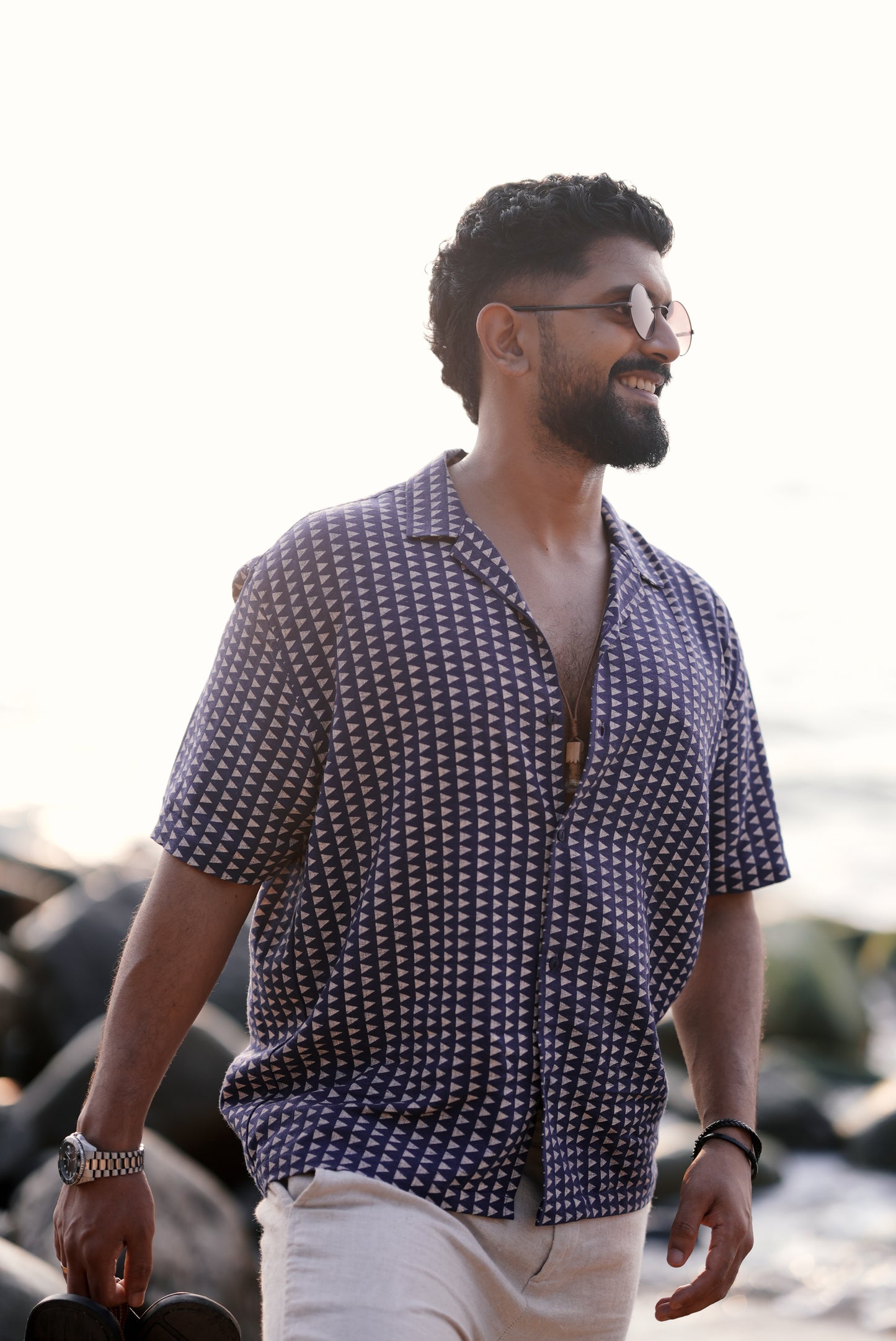 A man wearing a navy blue half-sleeve shirt with a white triangle print from Mod Mavens' new menswear collection. He pairs the shirt with light beige linen pants and a silver wristwatch, standing on a sandy beach lined with rocks, smiling and looking relaxed.
