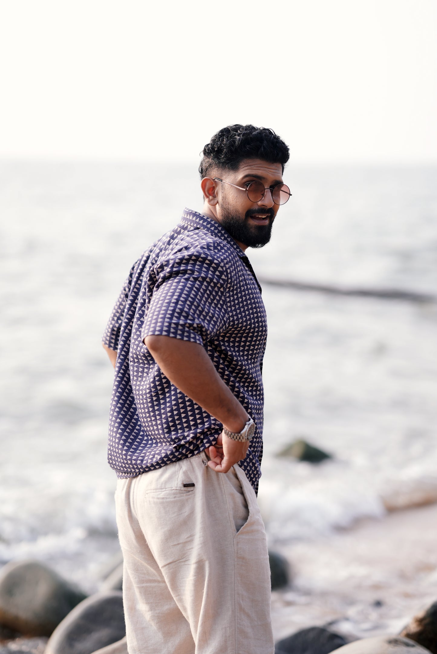 A man wearing a navy blue half-sleeve shirt with a white triangle print from Mod Mavens' new menswear collection. He pairs the shirt with light beige linen pants and a silver wristwatch, standing on a sandy beach lined with rocks, smiling and looking relaxed showing his side profile.