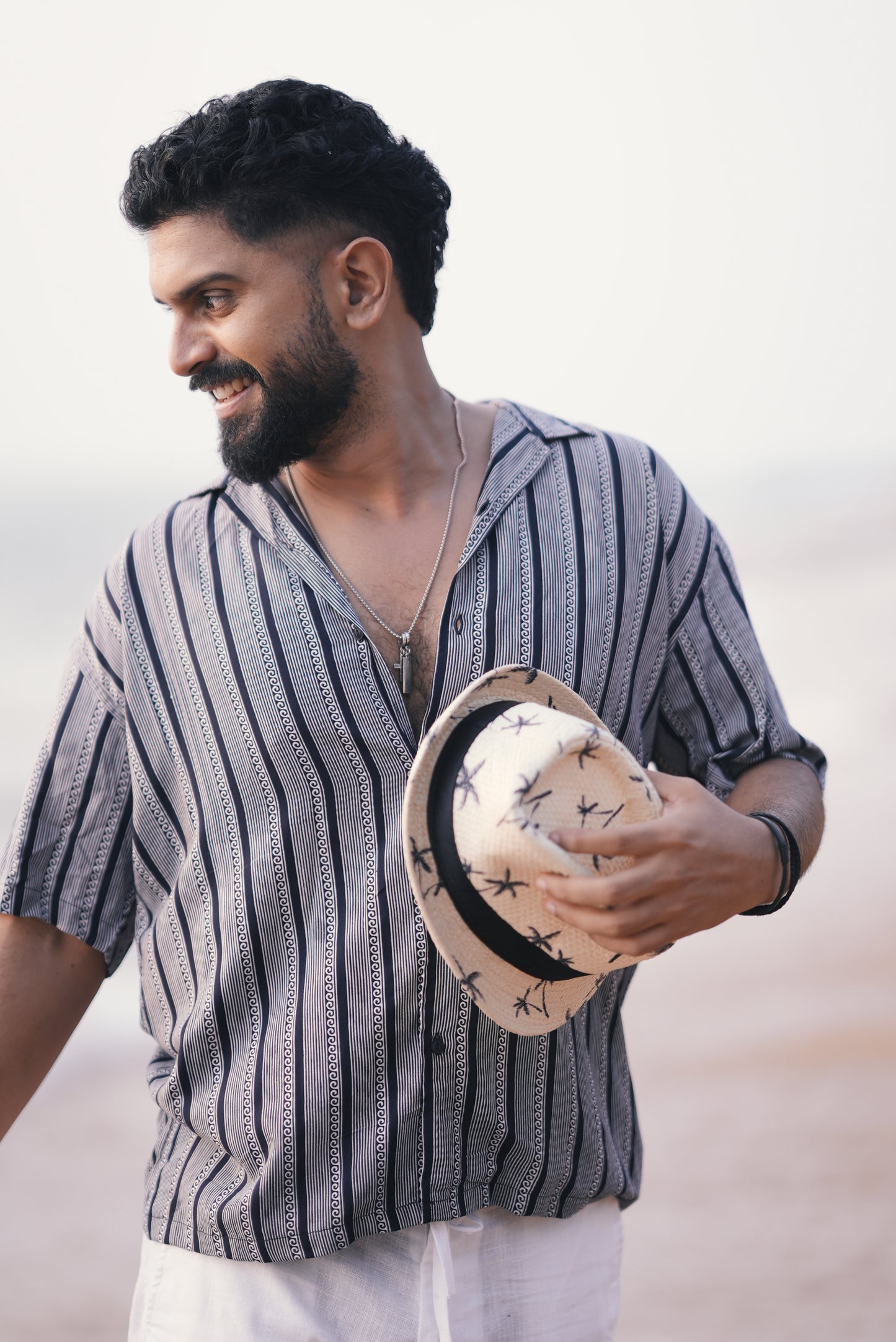 A man wearing a white and navy blue half-sleeve shirt with vertical wave and stripe patterns from Mod Mavens' menswear collection. He pairs the shirt with white linen trousers and accessorizes with layered necklaces and a black wristband. He is holding a straw hat with black palm tree prints, standing on a beach with a relaxed and cheerful expression.