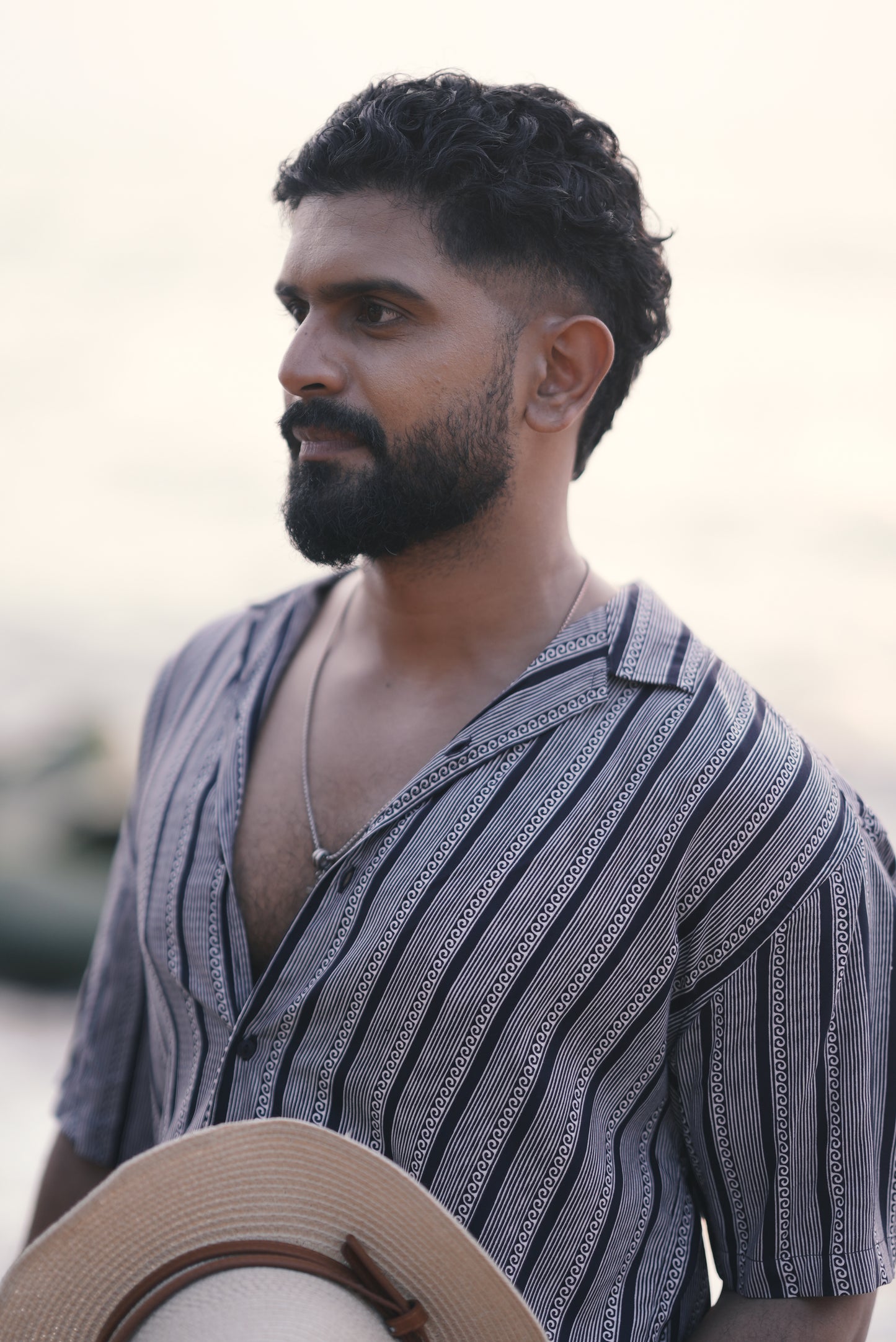 A man wearing a white and navy blue half-sleeve shirt with vertical wave and stripe patterns from Mod Mavens' menswear collection. He pairs the shirt with white linen trousers and accessorizes with layered necklaces and a black wristband. He is holding a straw hat with black palm tree prints, standing on a beach with a relaxed expression.