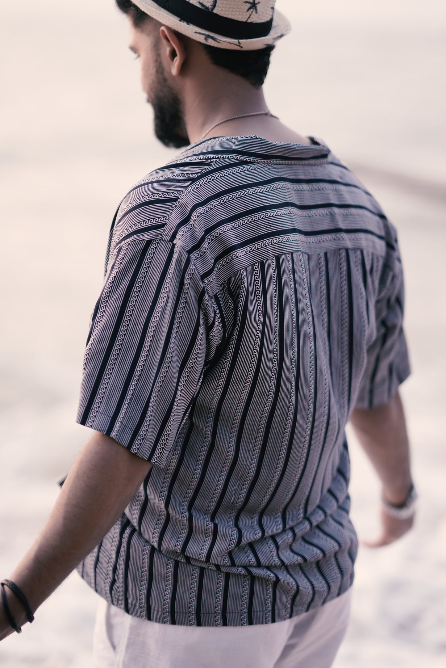 A side view of a man wearing a white and navy blue half-sleeve shirt with vertical wave and stripe patterns from Mod Mavens' menswear collection. The shirt's pattern and relaxed fit are highlighted as he stands on a sandy beach, creating an effortless and stylish look.