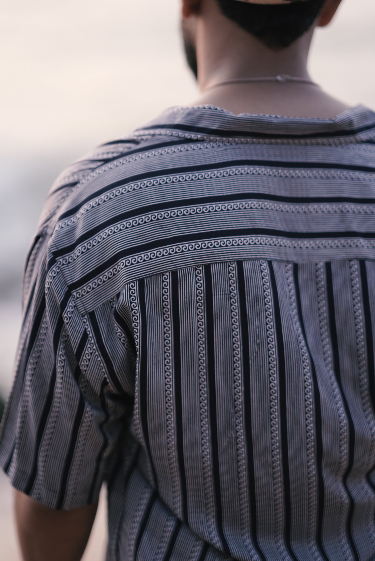 A back view of a man wearing a white and navy blue half-sleeve shirt with vertical wave and stripe patterns from Mod Mavens' menswear collection. The shirt's pattern and relaxed fit are highlighted as he stands on a sandy beach, creating an effortless and stylish look.
