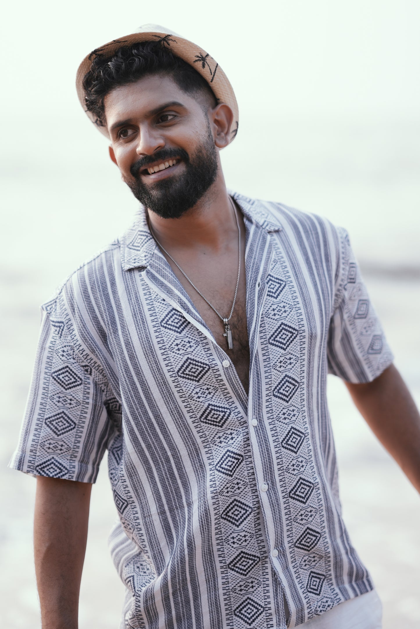 A model wearing a stylish short-sleeve button-up shirt with a geometric tribal weave pattern in shades of white and blue. The shirt has a casual, lightweight fabric, perfect for a beach or vacation vibe. He’s smiling, exuding relaxed energy.