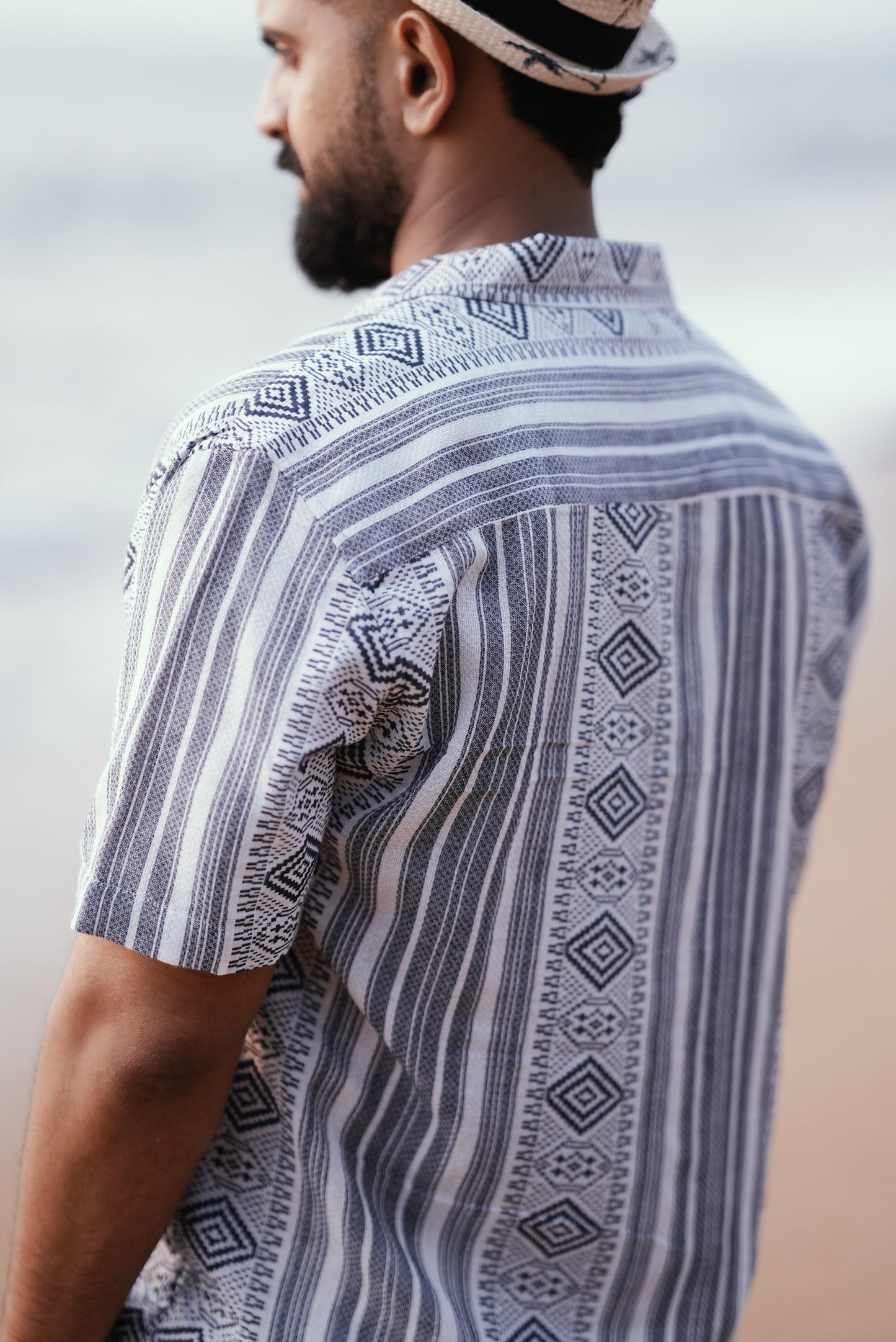 A back view model wearing a stylish short-sleeve button-up shirt with a geometric tribal weave pattern in shades of white and blue. The shirt has a casual, lightweight fabric, perfect for a beach or vacation vibe.