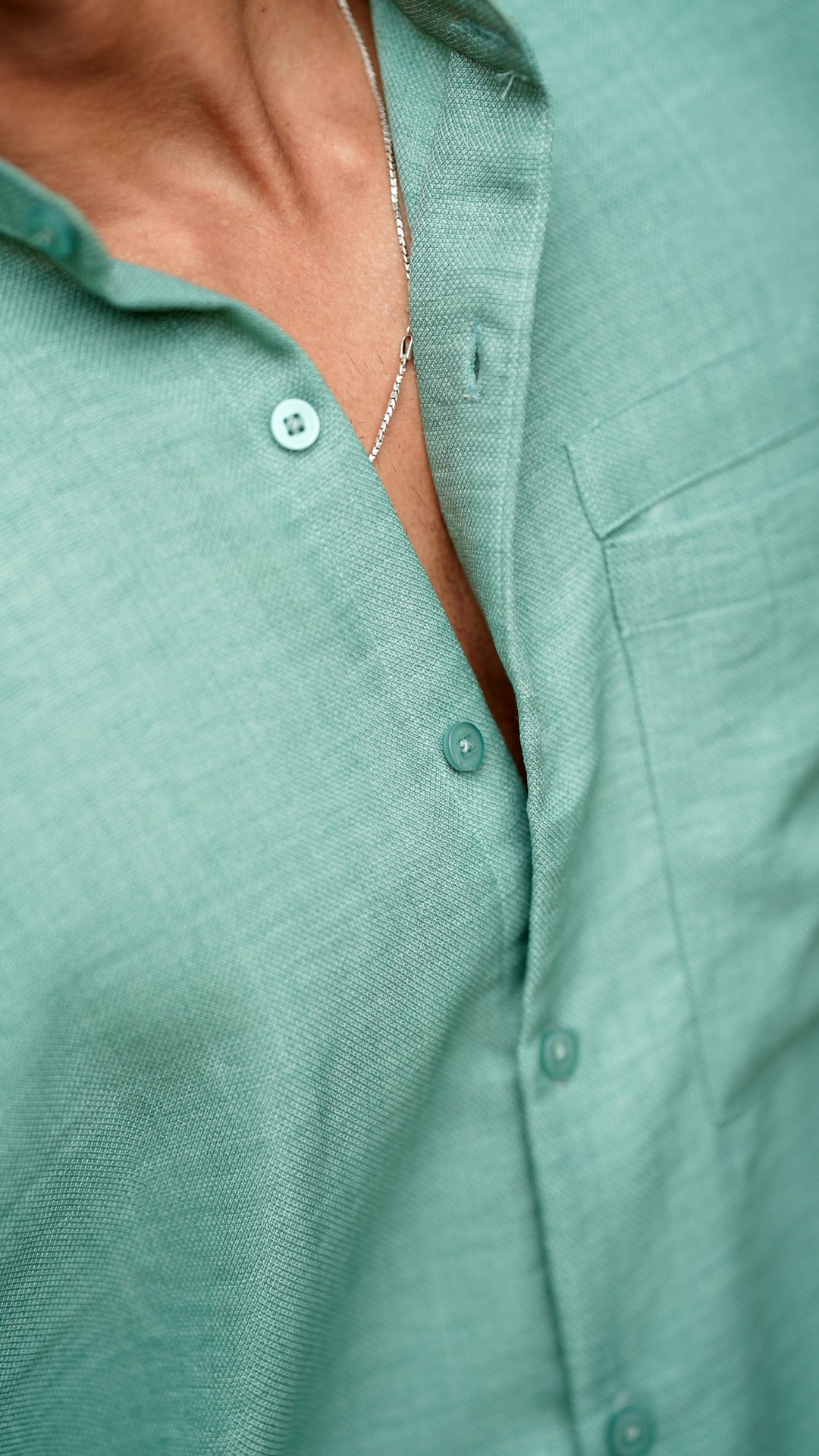 Close-up view of a male model wearing a pastel green, slim-fit, full-sleeve shirt from Mod Maven. The shirt is buttoned down with matching green buttons, showing a sleek, minimalistic design. He is accessorized with a thin silver chain, adding a touch of elegance to the outfit.
