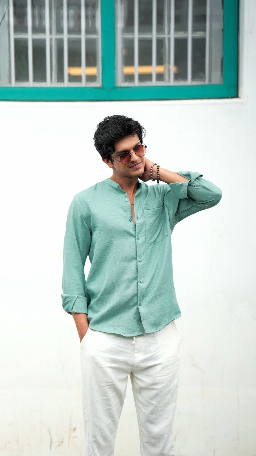 A male model wearing a pastel green, slim-fit, full-sleeve shirt from Mod Maven. He has short, tousled black hair, and wears an off white linen trousers and round, tinted sunglasses. The shirt has a soft texture with matching buttons. He is standing in front of a cafe wall, exuding a casual yet refined look.