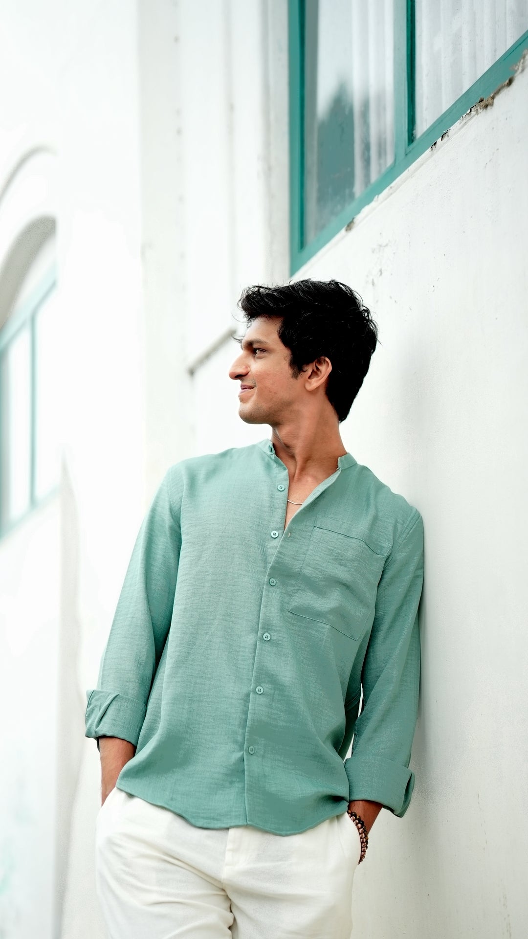 A male model wearing a pastel green, slim-fit, full-sleeve shirt from Mod Maven. He has short, tousled black hair, and wears an off white linen trousers and round, tinted sunglasses. The shirt has a soft texture with matching buttons. He is standing in front of a cafe wall, exuding a casual yet refined look.