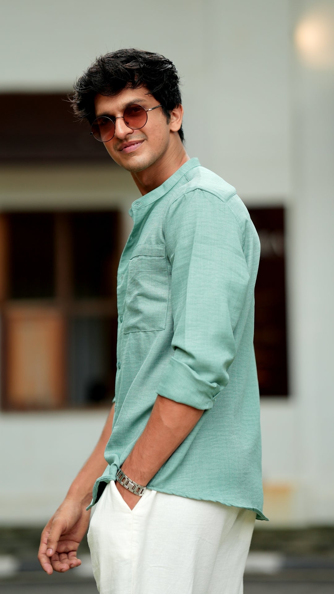 Side look of a male model wearing a pastel green, slim-fit, full-sleeve shirt from Mod Maven. He has short, tousled black hair, and wears round, tinted sunglasses. The shirt has a soft texture with matching green buttons. He is standing in front of a neutral-toned cafe wall, exuding a casual yet refined look.