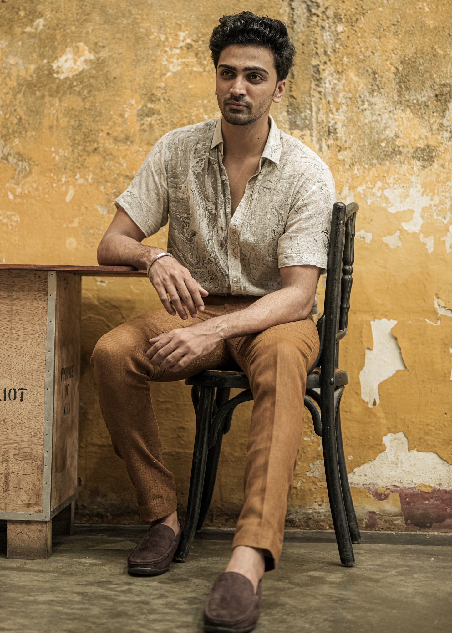 Mod Maven's stylish black and white marble-printed half sleeve shirt, made from breathable dobby cotton, is showcased in this image. The model sits in a unique cafe with a rough, broken wall theme, adorned in the fashionable shirt paired with formal mud brown trousers. The yellow wall paint of the cafe adds a vibrant contrast, creating a visually appealing and trendy atmosphere.