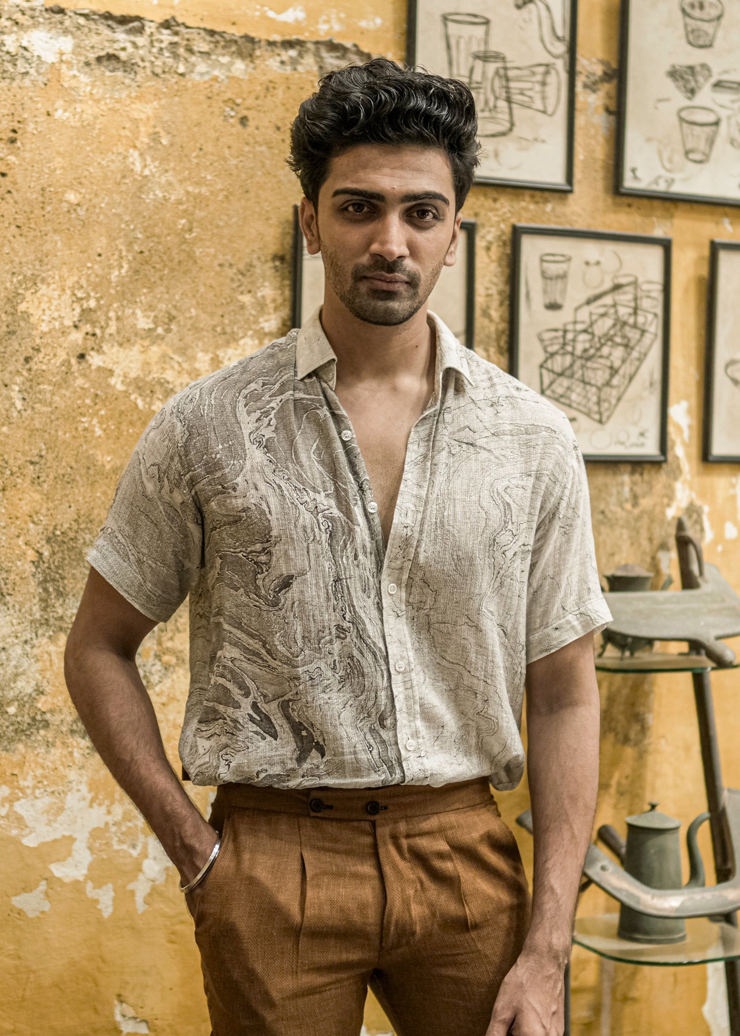 A model standing confidently in a unique cafe with a rough, broken wall theme, featuring vibrant yellow wall paint. The model is wearing a stylish black and white marble-printed half-sleeve shirt from the brand Mod Maven, made of dobby cotton. Paired with the shirt is a set of formal mud-brown trousers, creating a fashionable and contemporary ensemble.
