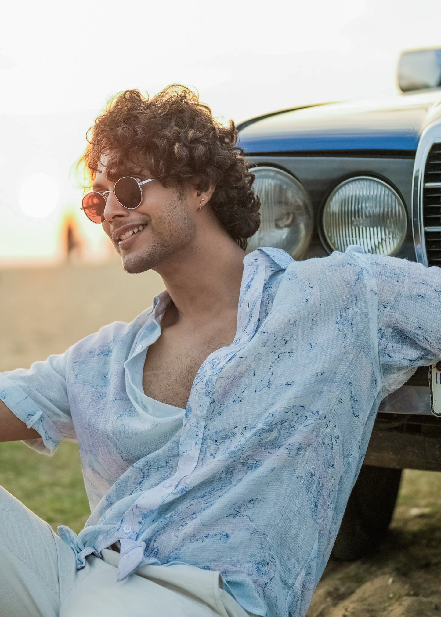 A relaxed beach scene featuring a model sitting on soft sand next to a blue vintage car. The model is wearing a stylish Dobby cotton marble printed half-sleeve shirt in shades of blue and lilac from the Mod Maven brand. The outfit is completed with off-white comfortable fit trousers, creating a casual and fashionable beach look.