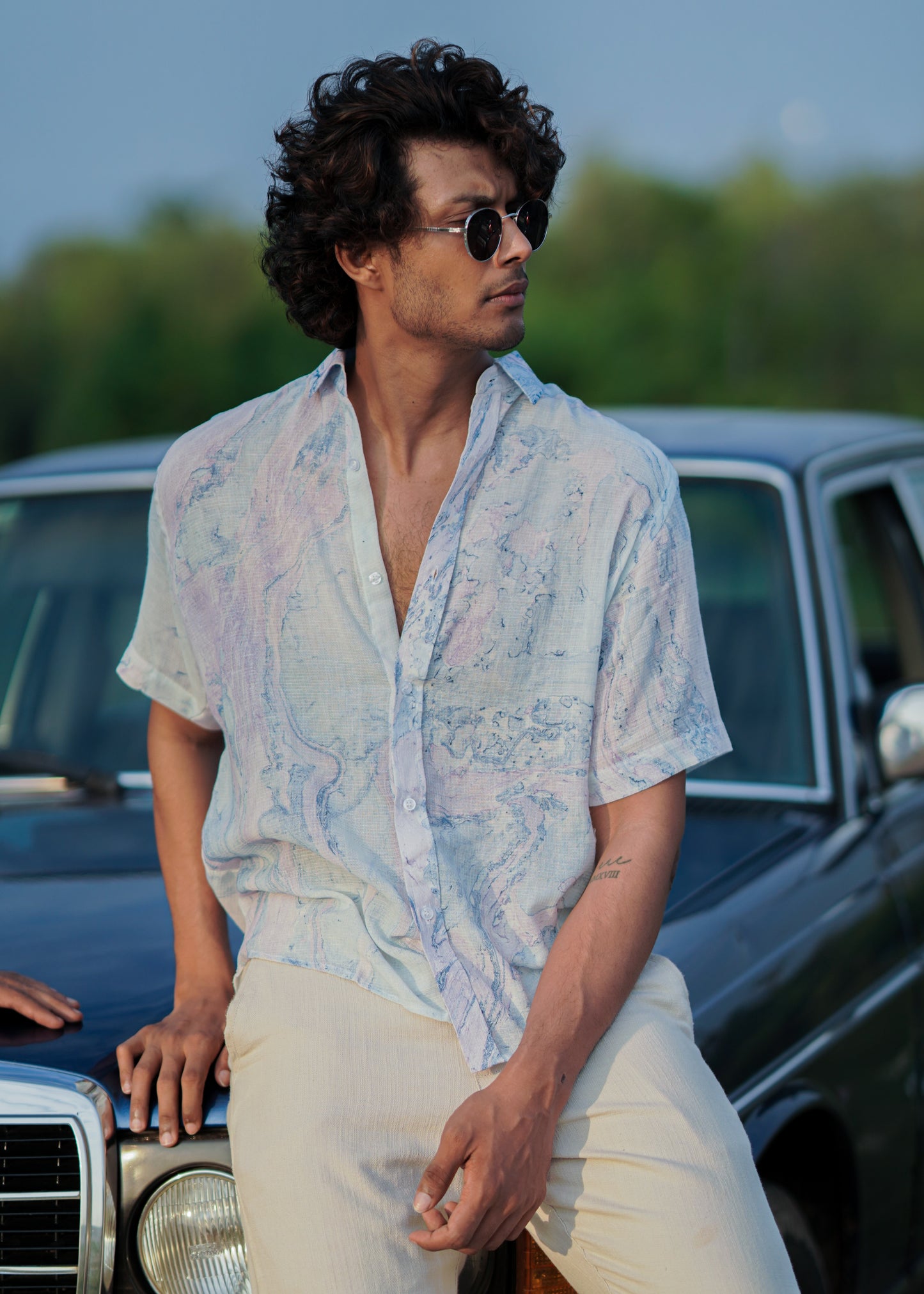 A relaxed beach scene featuring the male model sitting on a blue vintage car. The model is wearing a stylish Dobby cotton marble printed half-sleeve shirt in shades of blue and lilac from the Mod Maven brand. The outfit is completed with off-white comfortable fit trousers and sunglasses creating a casual and fashionable beach look.