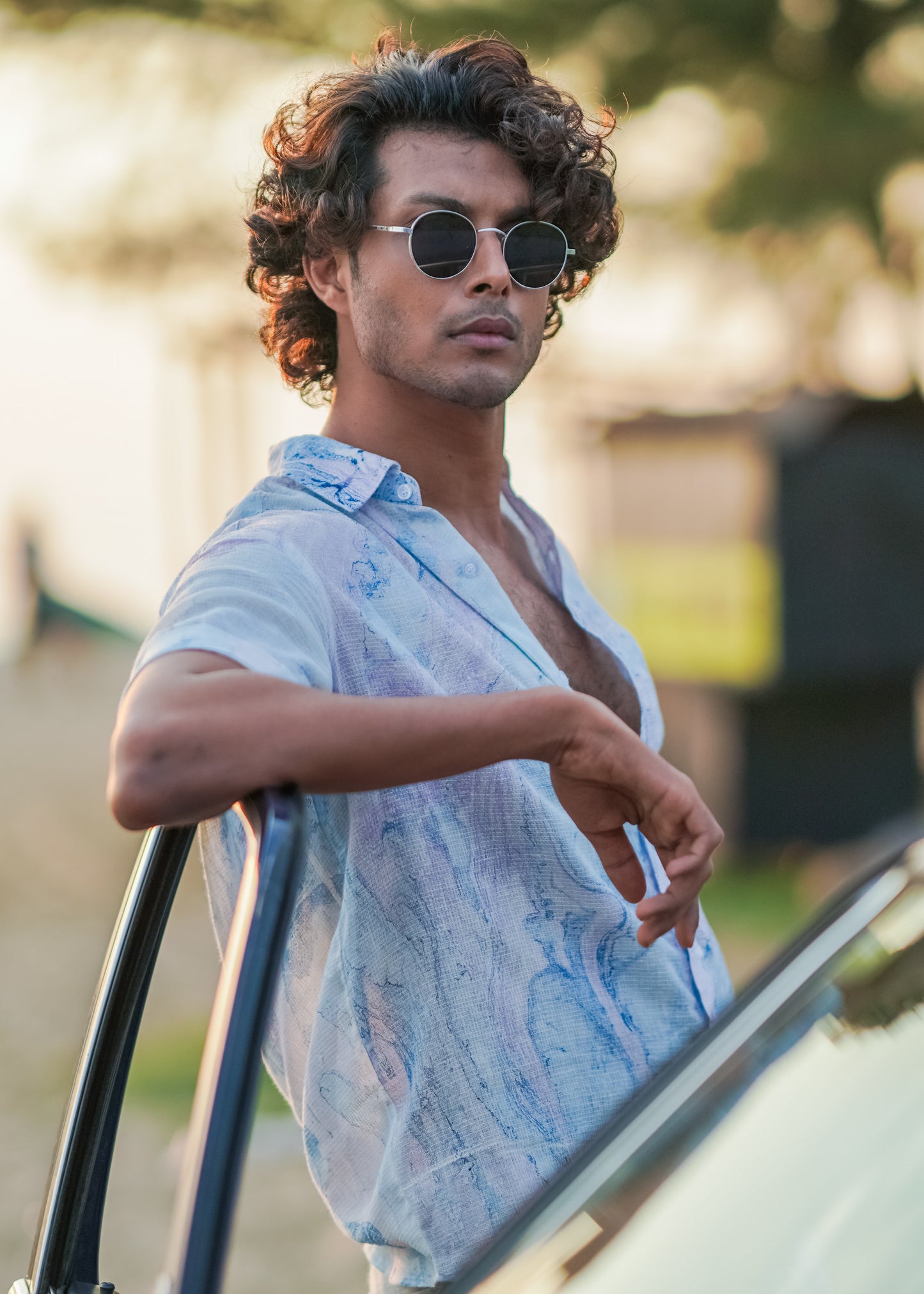 A relaxed beach scene featuring the male model leaning on the opened front door of a blue vintage car. The model is wearing a stylish Dobby cotton marble printed half-sleeve shirt in shades of blue and lilac from the Mod Maven brand. The outfit is completed with off-white comfortable fit trousers and sunglasses creating a casual and fashionable beach look.