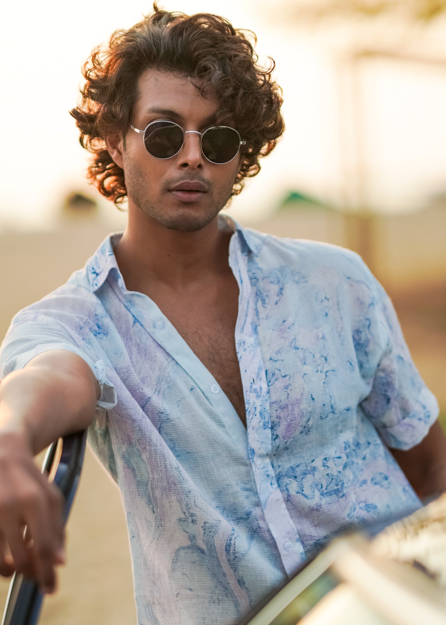 A relaxed beach scene featuring the male model leaning on the opened front door of a blue vintage car. The model is wearing a stylish Dobby cotton marble printed half-sleeve shirt in shades of blue and lilac from the Mod Maven brand. The outfit is completed with off-white comfortable fit trousers and sunglasses creating a casual and fashionable beach look.
