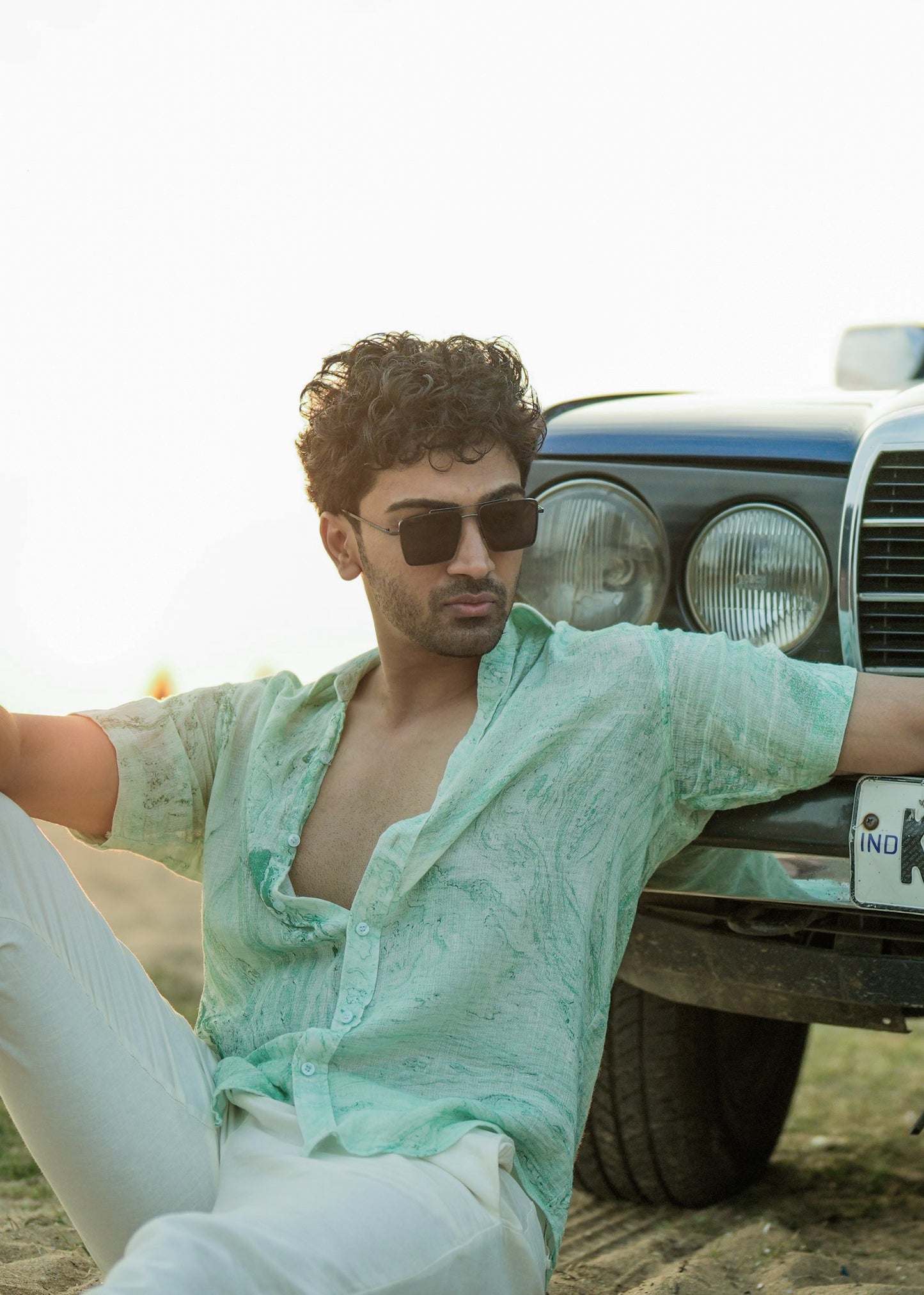 An image featuring a model seated on beach sand, leaning against a vibrant blue vintage car. The model is wearing a stylish Green Dobby Cotton Marble Printed Half Sleeve Shirt from the brand Mod Maven. The shirt complements the beachy vibes with its unique pattern. Paired with the shirt, the model sports off-white comfortable fit trousers, creating a relaxed yet fashionable beach ensemble.