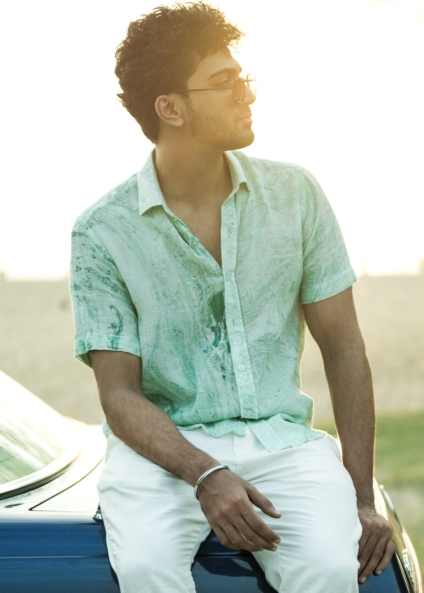 An image featuring a model seated on a vibrant blue vintage car in a beach. The model is wearing a stylish Green Dobby Cotton Marble Printed Half Sleeve Shirt from the brand Mod Maven. The shirt complements the beachy vibes with its unique pattern. Paired with the shirt, the model sports off-white comfortable fit trousers, creating a relaxed yet fashionable beach ensemble.