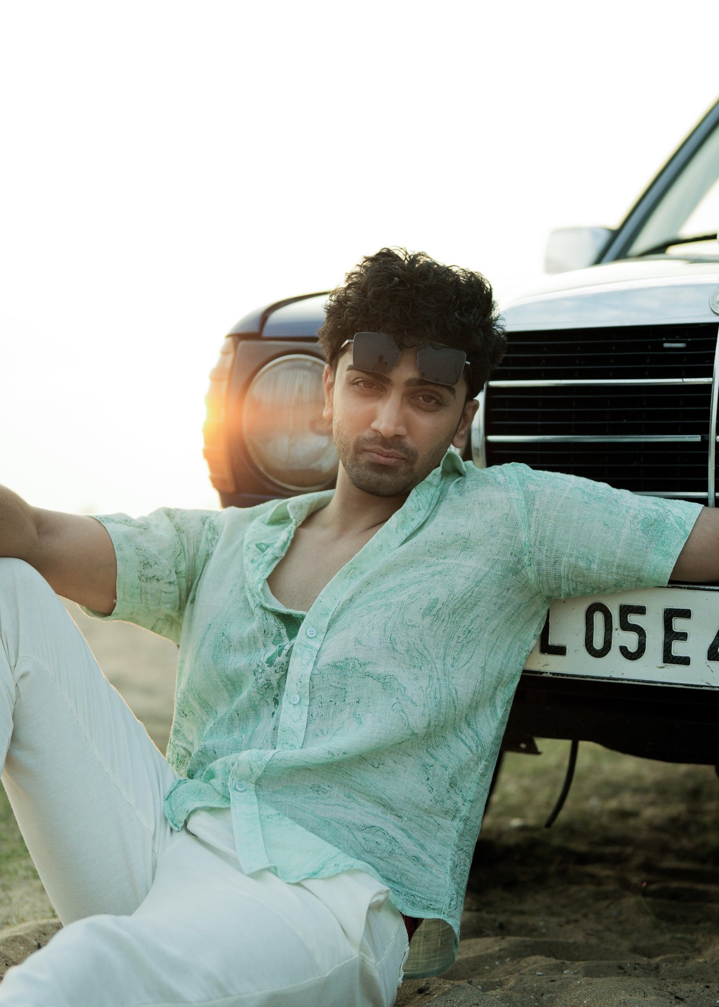 An image featuring a model seated on beach sand, leaning against a vibrant blue vintage car. The model is wearing a stylish Green Dobby Cotton Marble Printed Half Sleeve Shirt from the brand Mod Maven. The shirt complements the beachy vibes with its unique pattern. Paired with the shirt, the model sports off-white comfortable fit trousers, creating a relaxed yet fashionable beach ensemble.