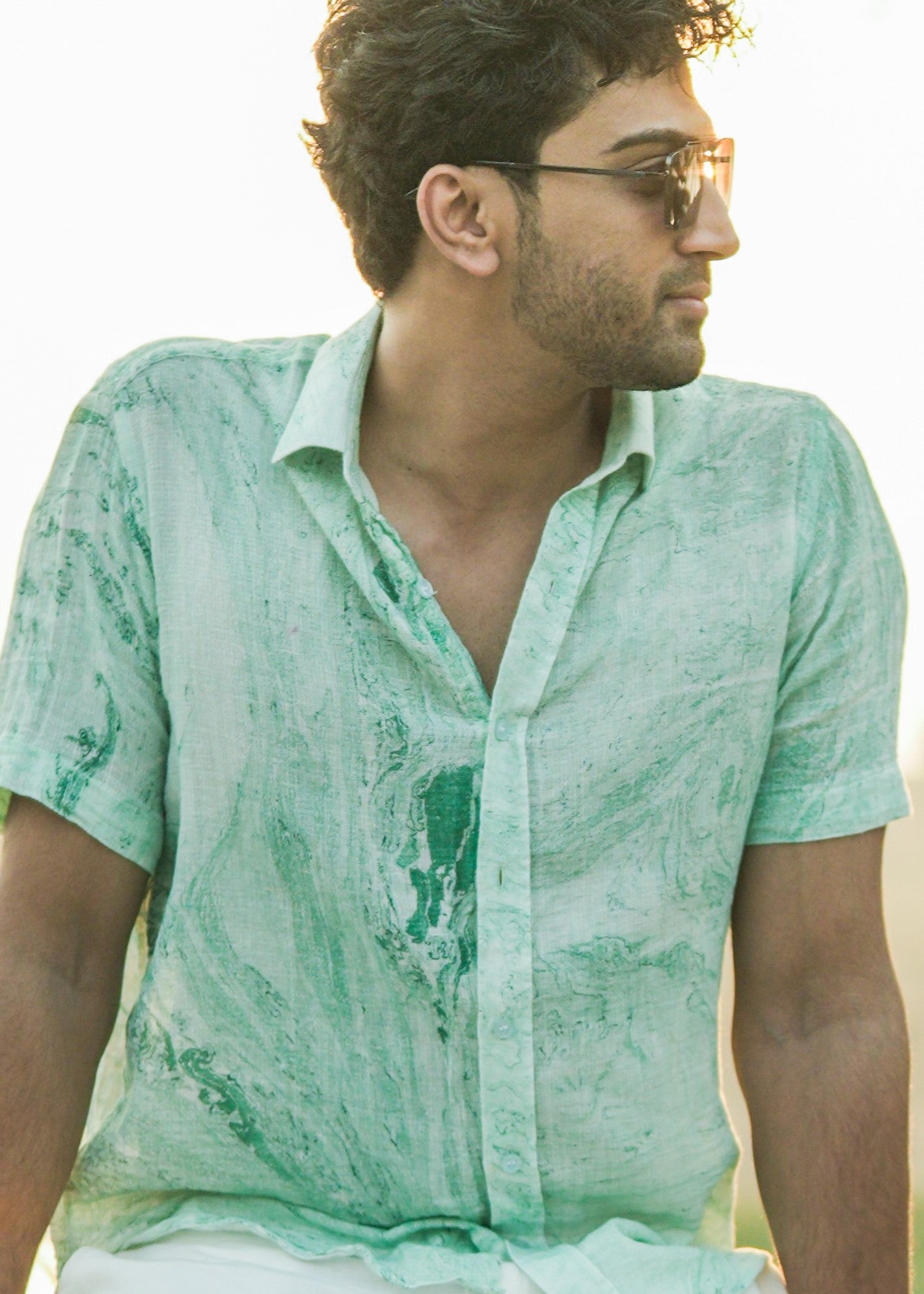 An image featuring a close up shot of the model seated, wearing a stylish Green Dobby Cotton Marble Printed Half Sleeve Shirt from the brand Mod Maven. The shirt complements the beachy vibes with its unique pattern paired with sunglasses creating a relaxed yet fashionable beach ensemble.