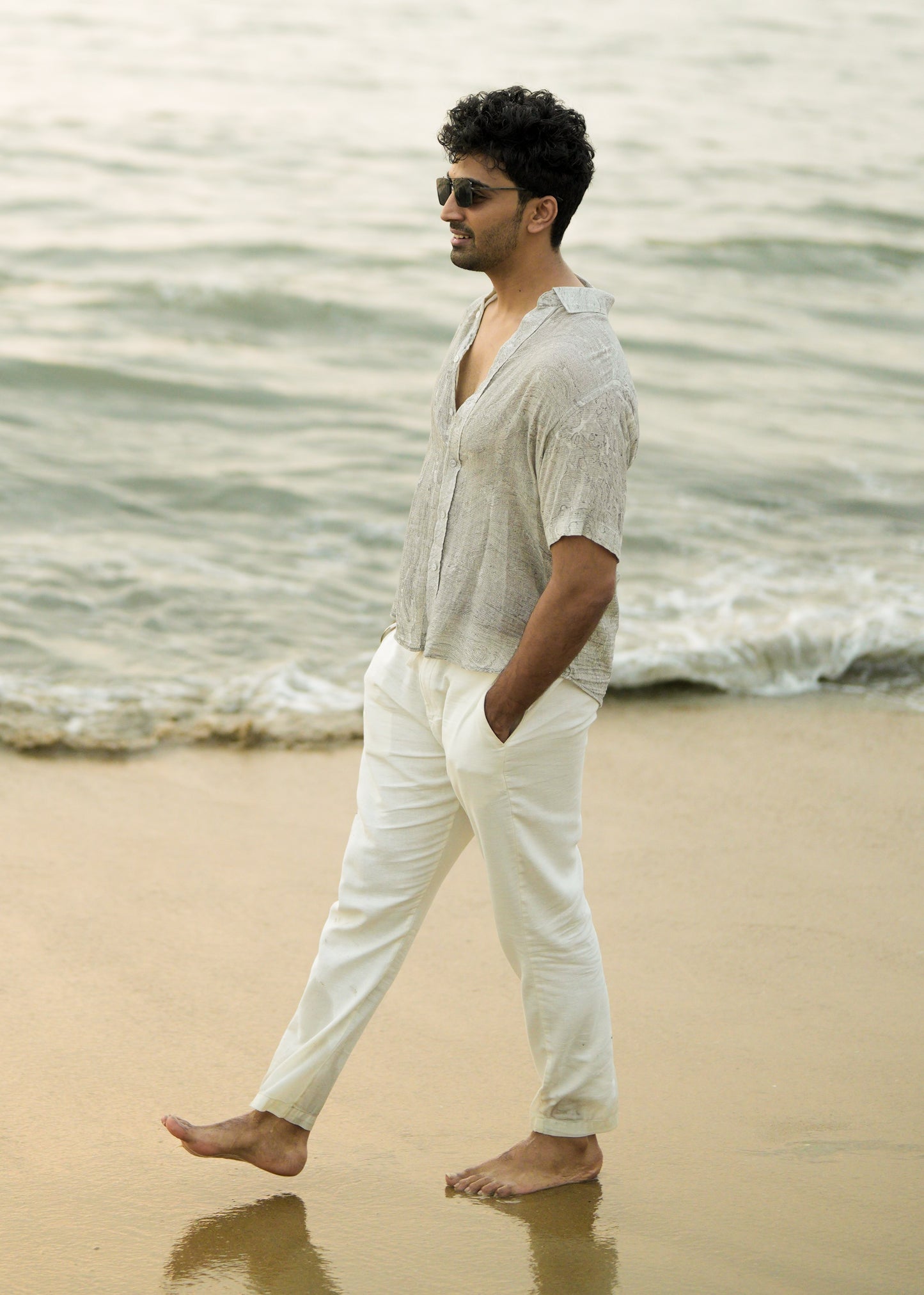 Male model strolling confidently along the beach, showcasing the effortlessly stylish look with a Mod Maven grey Dobby cotton marble printed half-sleeve shirt. The shirt complements off-white comfortable fit trousers, creating a chic and relaxed ensemble. The model completes the look with trendy sunglasses, embodying the laid-back elegance of the Mod Maven brand.