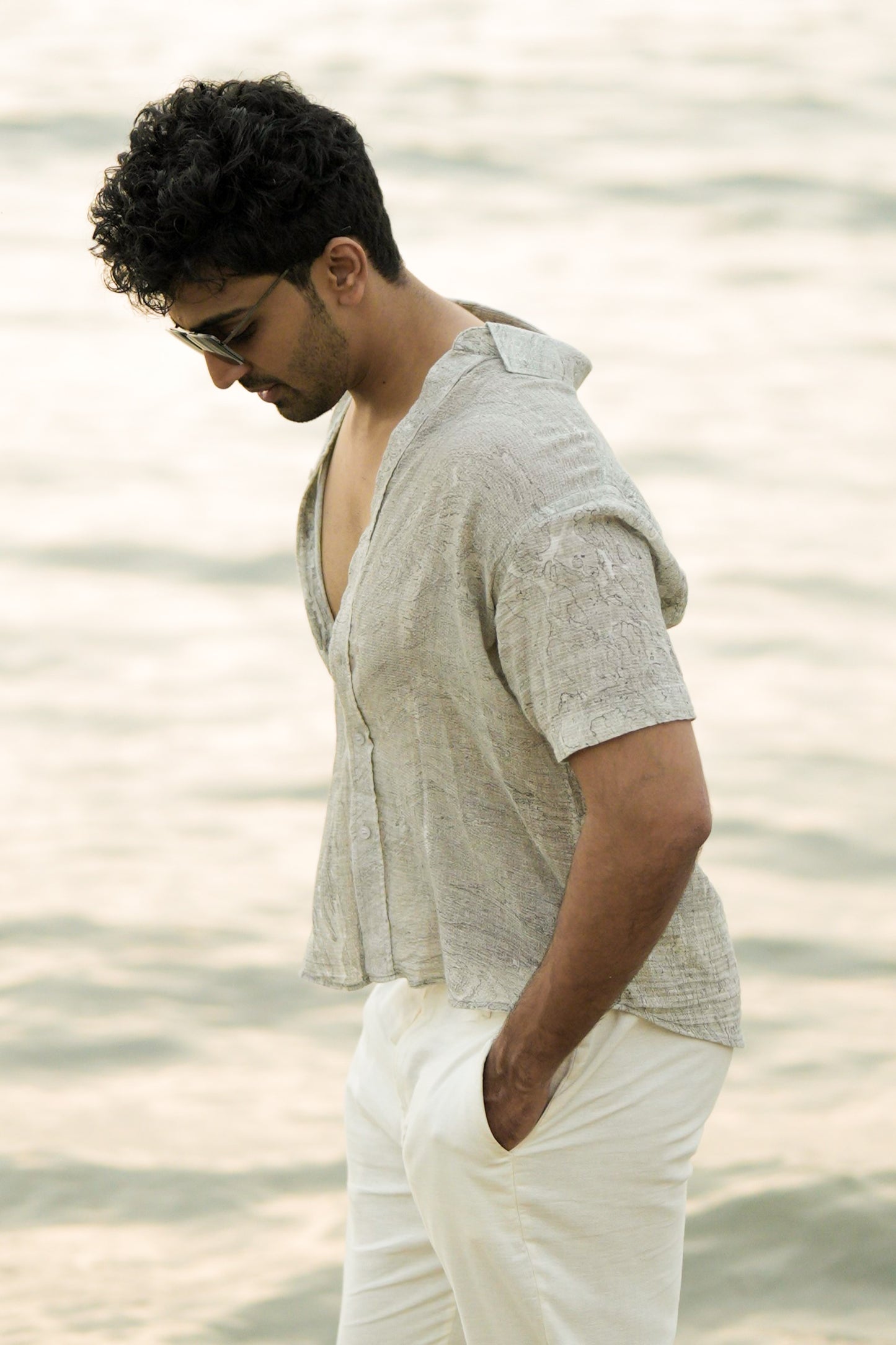 Close up shot of Male model strolling confidently along the beach, showcasing the effortlessly stylish look with a Mod Maven grey Dobby cotton marble printed half-sleeve shirt. The shirt complements off-white comfortable fit trousers, creating a chic and relaxed ensemble. The model completes the look with trendy sunglasses, embodying the laid-back elegance of the Mod Maven brand.
