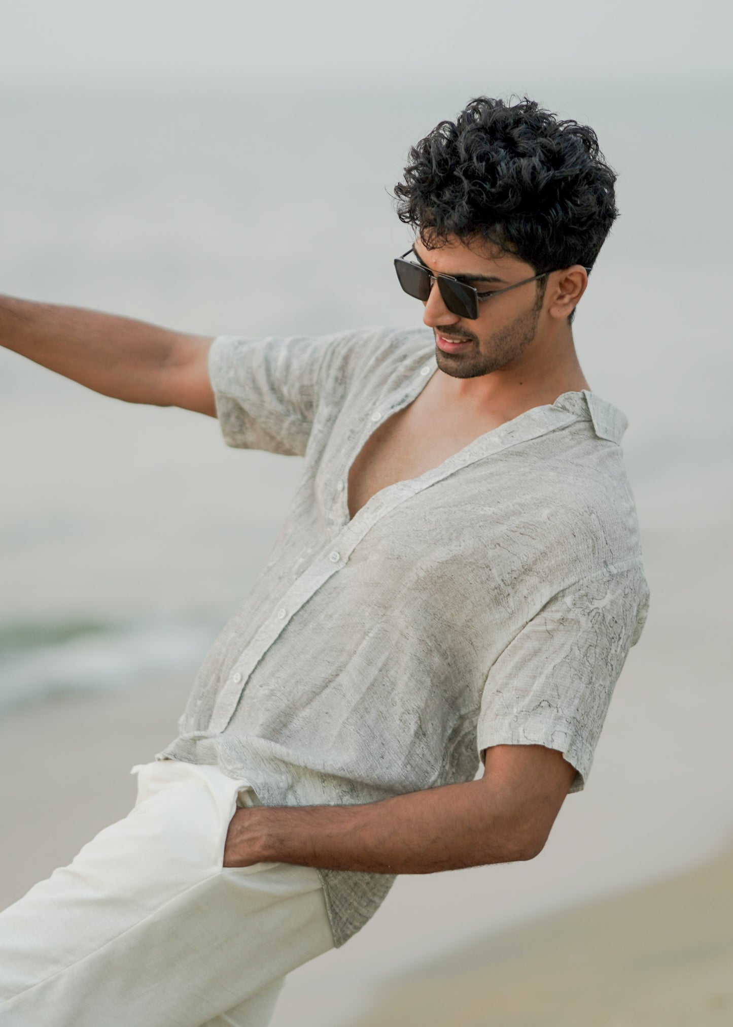 Male model strolling confidently along the beach, showcasing the effortlessly stylish look with a Mod Maven grey Dobby cotton marble printed half-sleeve shirt. The shirt complements off-white comfortable fit trousers, creating a chic and relaxed ensemble. The model completes the look with trendy sunglasses, embodying the laid-back elegance of the Mod Maven brand.