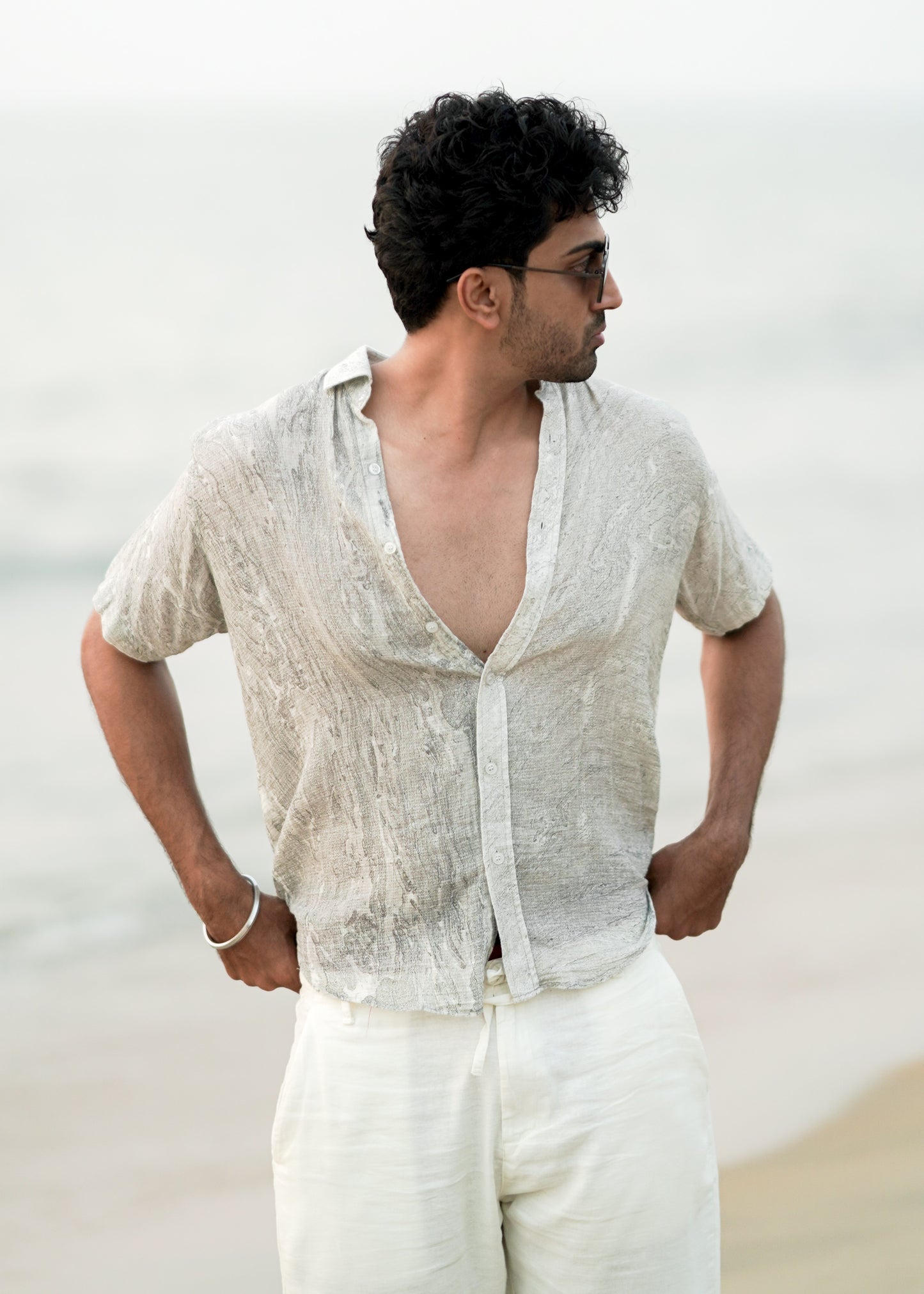 Male model strolling confidently along the beach, showcasing the effortlessly stylish look with a Mod Maven grey Dobby cotton marble printed half-sleeve shirt. The shirt complements off-white comfortable fit trousers, creating a chic and relaxed ensemble. The model completes the look with trendy sunglasses, embodying the laid-back elegance of the Mod Maven brand.