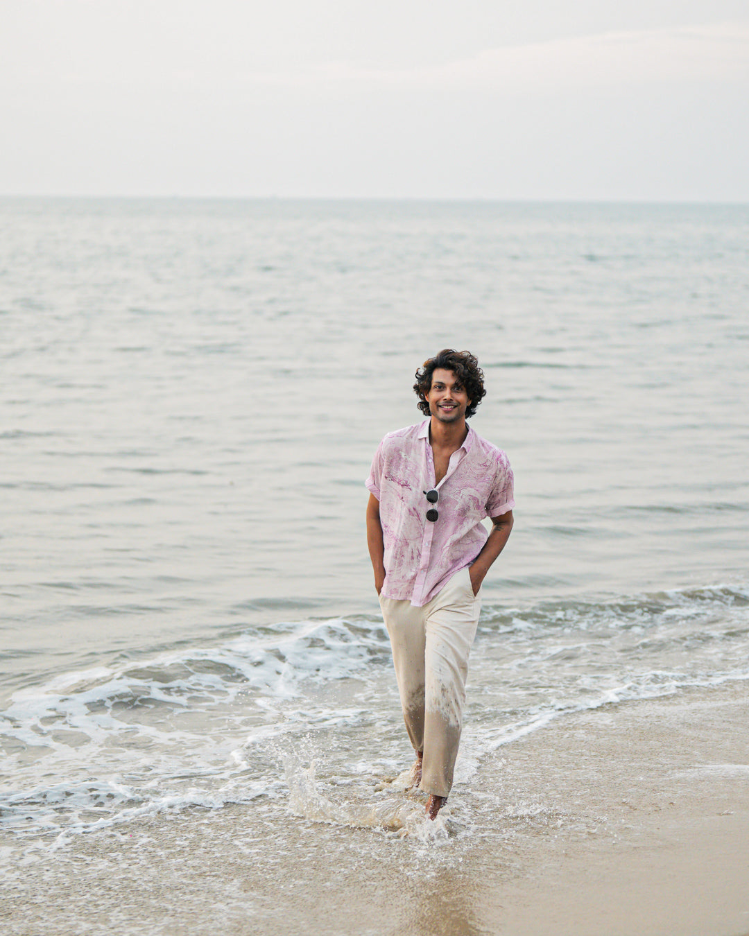 An image featuring a male model walking on the beach, showcasing the effortless style of Mod Maven's dobby cotton marble printed half sleeve shirt in a soothing lilac color. Paired with off-white comfortable fit trousers, the outfit exudes a casual yet refined vibe. The model wears sunglasses casually hung on the shirt, adding a touch of beach-ready sophistication to this coastal-inspired look.
