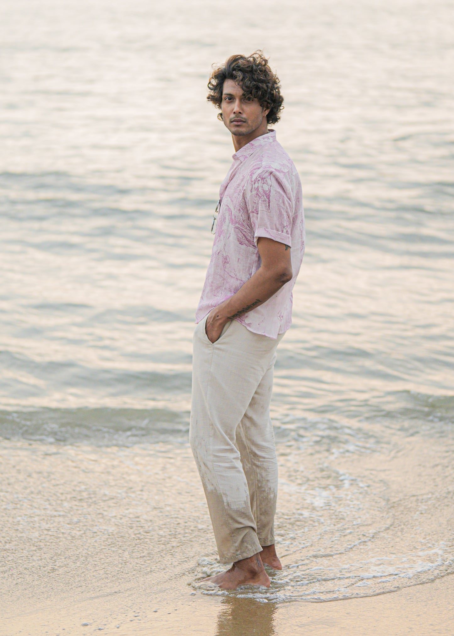An image featuring side profile of a male model walking on the beach, showcasing the effortless style of Mod Maven's dobby cotton marble printed half sleeve shirt in a soothing lilac color. Paired with off-white comfortable fit trousers, the outfit exudes a casual yet refined vibe. The model wears sunglasses casually hung on the shirt, adding a touch of beach-ready sophistication to this coastal-inspired look."