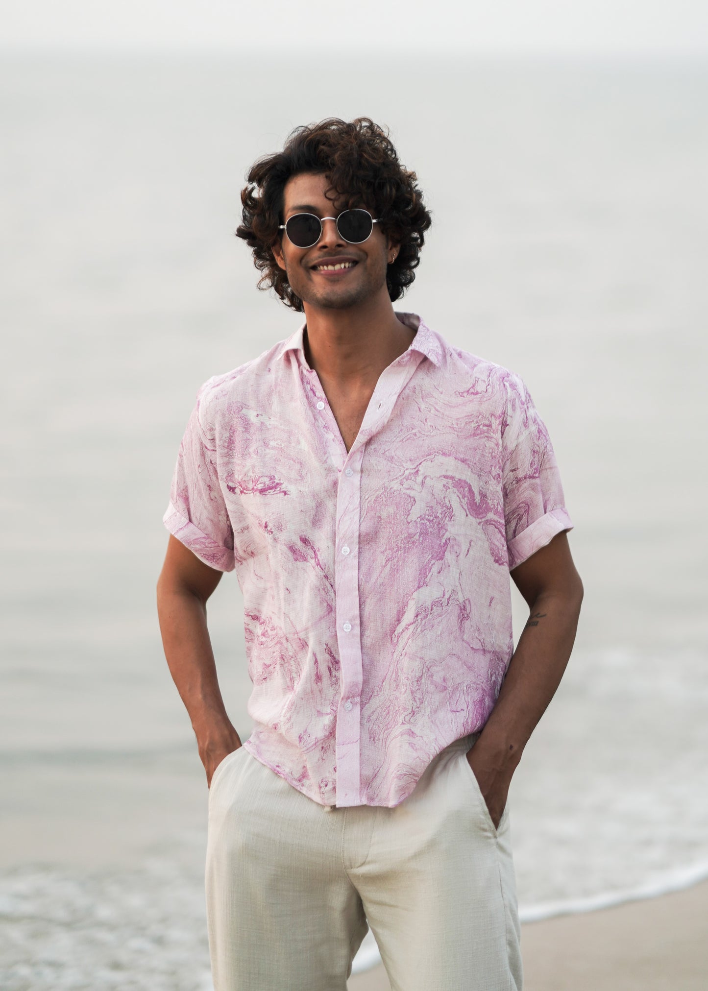 An image featuring a male model standing on the beach, showcasing the effortless style of Mod Maven's dobby cotton marble printed half sleeve shirt in a soothing lilac color. Paired with off-white comfortable fit trousers, the outfit exudes a casual yet refined vibe. The model wears sunglasses casually, adding a touch of beach-ready sophistication to this coastal-inspired look.