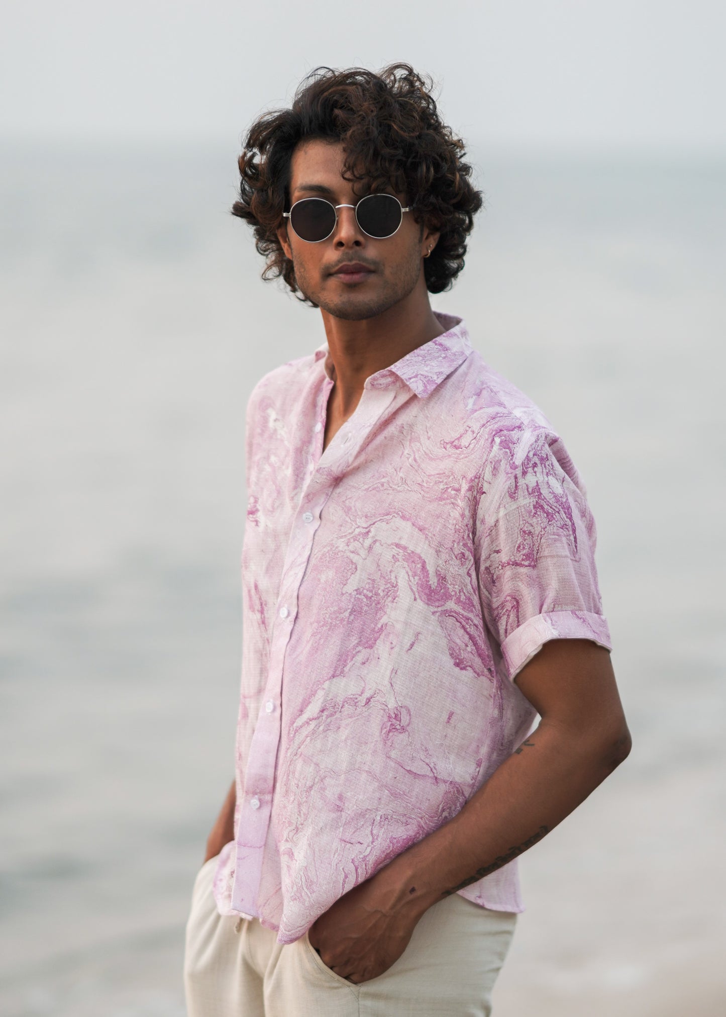 Side profile Image featuring a male model walking on the beach, showcasing the effortless style of Mod Maven's dobby cotton marble printed half sleeve shirt in a soothing lilac color. Paired with off-white comfortable fit trousers, the outfit exudes a casual yet refined vibe. The model wears sunglasses casually hung on the shirt, adding a touch of beach-ready sophistication to this coastal-inspired look.