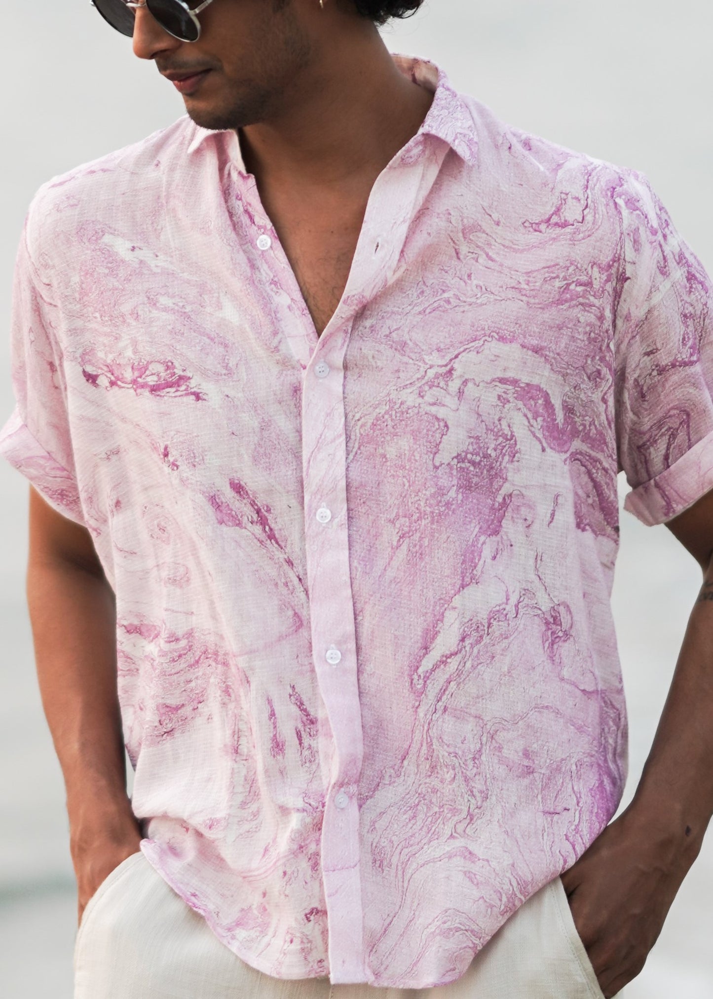 "An image featuring a male model walking on the beach, showcasing effortless style in a lilac-colored, dobby cotton marble printed half-sleeve shirt from the Mod Maven brand. Paired with comfortable off-white trousers, the ensemble exudes a relaxed vibe. The focus is on the close-up shot of the shirt's texture and pattern, revealing intricate details that add a touch of sophistication.