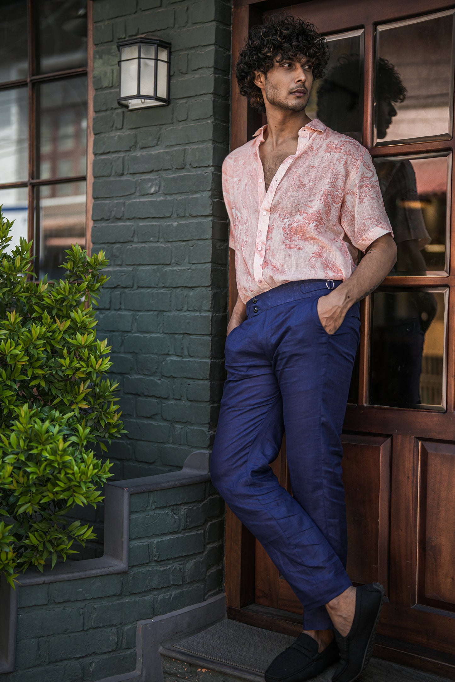 A stylish male model stands confidently in front of a dark brown wooden cafe door with a military green brick wall backdrop. He is wearing a fashion-forward Dobby cotton marble printed half-sleeve shirt in vibrant orange from the brand Mod Maven. The shirt is perfectly paired with navy blue formal fit trousers, creating a sophisticated and trendy ensemble.