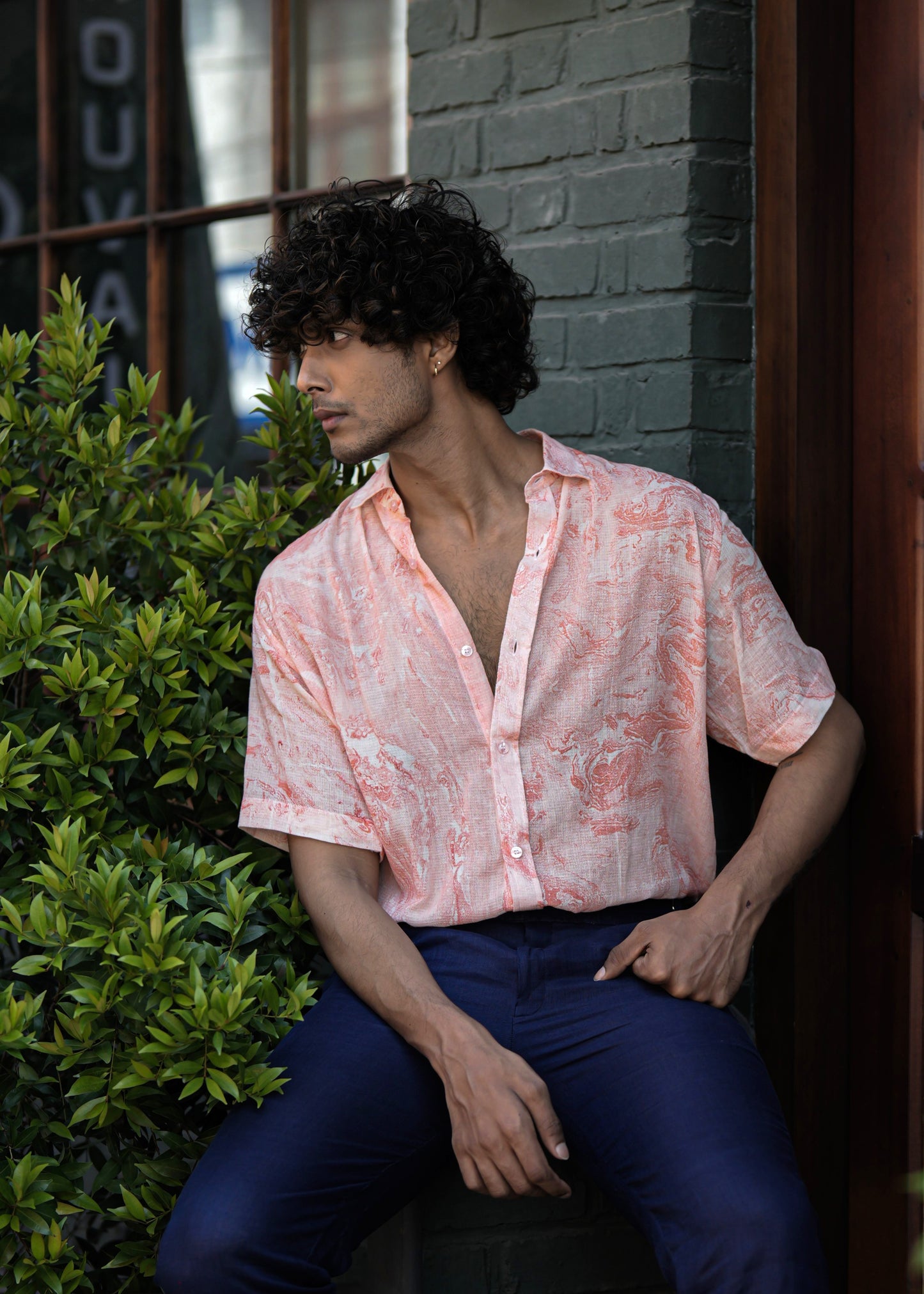 Male model sitting in front of a dark brown wood cafe door, set against a military green brick wall. The model wears a stylish Dobby cotton marble printed half-sleeve shirt in vibrant orange from the Mod Maven brand, paired with navy blue formal fit trousers.